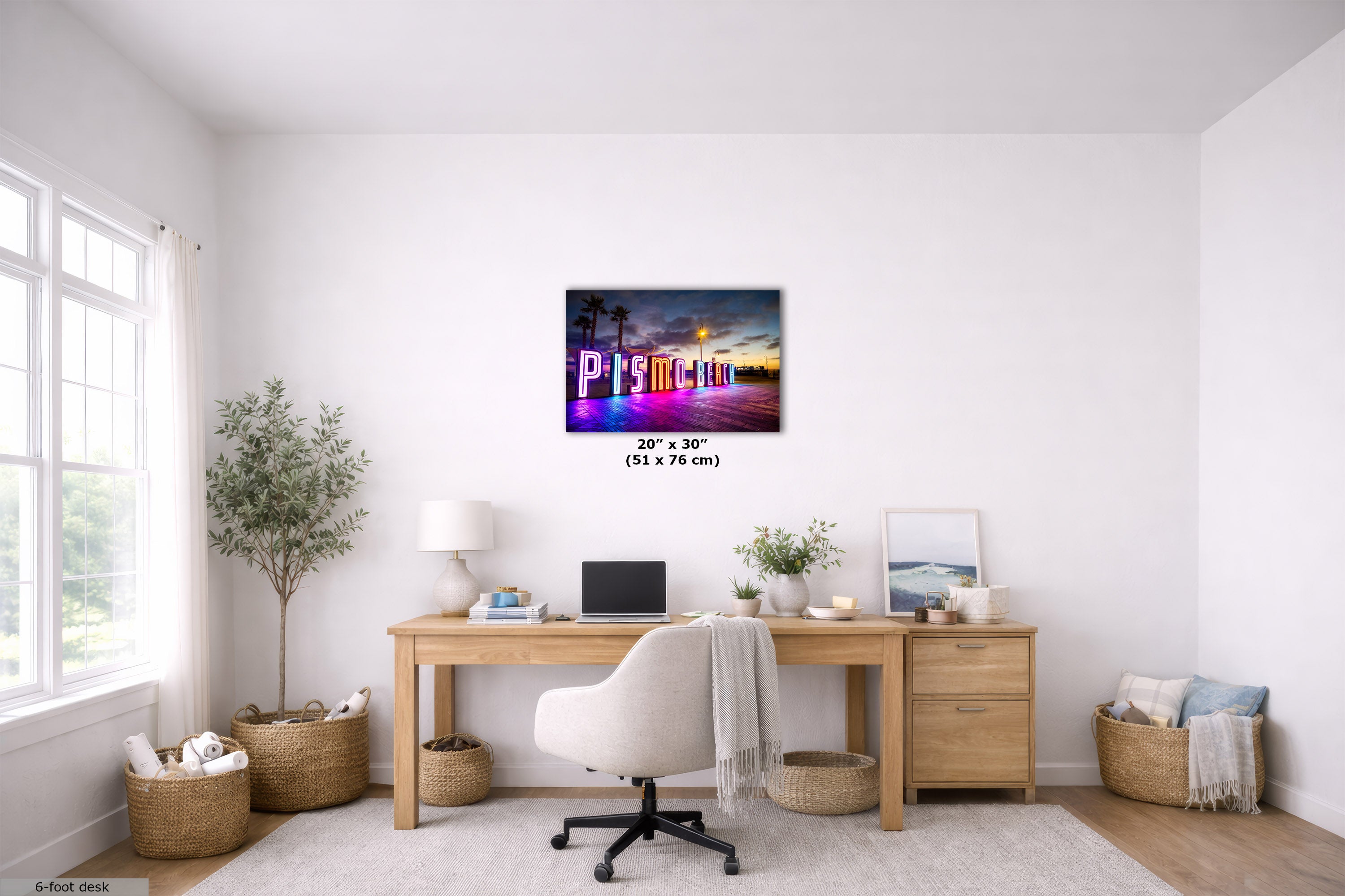 A 20x30 inch canvas print featuring a colorful sunset beach scene with the word 'Pismo Beach' in large letters, displayed on a white wall in a modern, bright home office setting above a wooden desk.