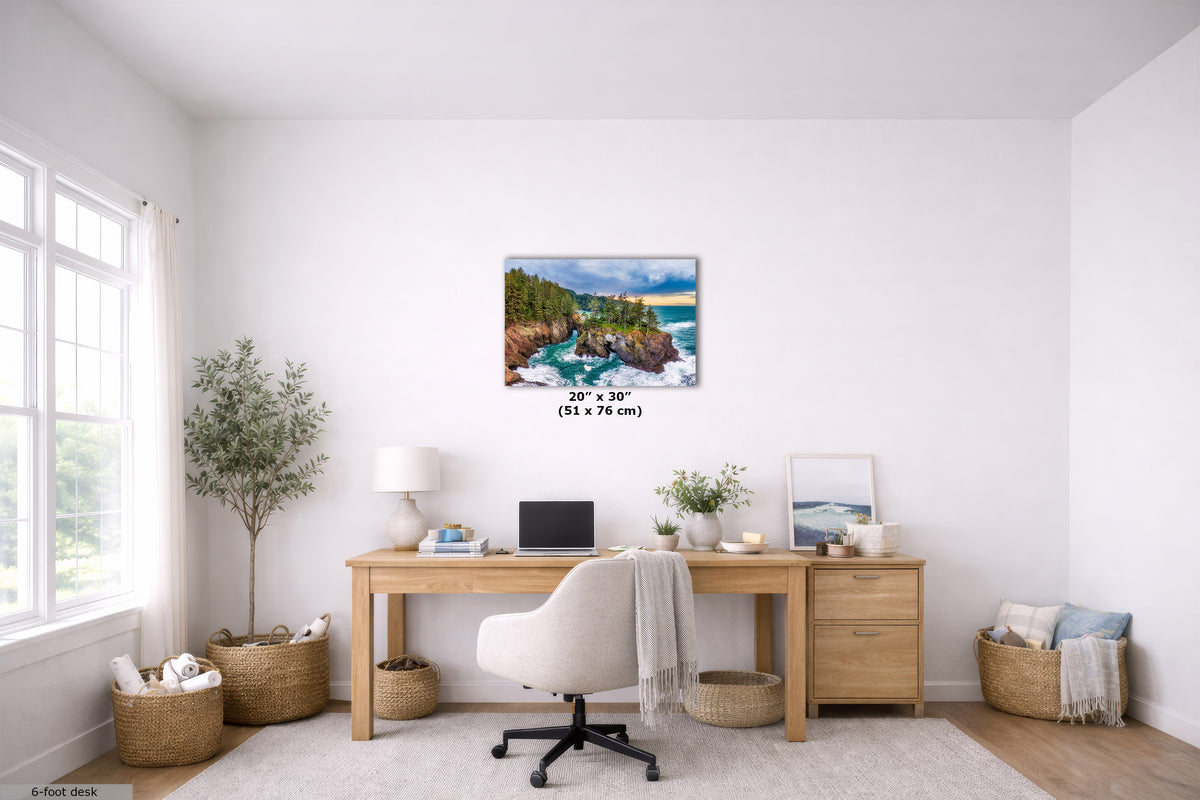 A coastal landscape canvas print displayed on a white wall in a bright, modern home office setting. The print features a rocky coastline with green trees, blue water, and a yellow-orange sunset sky.