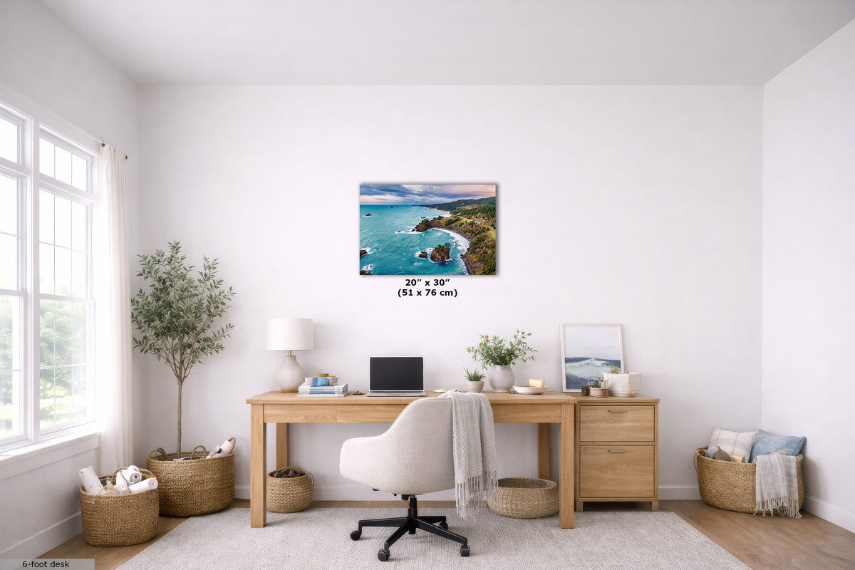A 20" x 30" square canvas print of a coastal rocky shoreline with turquoise water, displayed on a white wall in a bright, modern home office setting above a wooden desk.