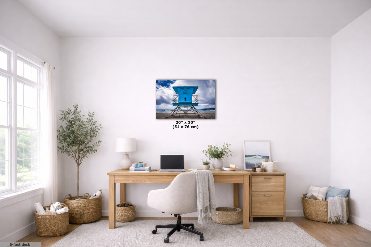 A wall art print showing a blue lifeguard tower over water, displayed above a wooden desk in a bright, modern home office. The print includes size text indicating 20" x 30" (51 x 76 cm).