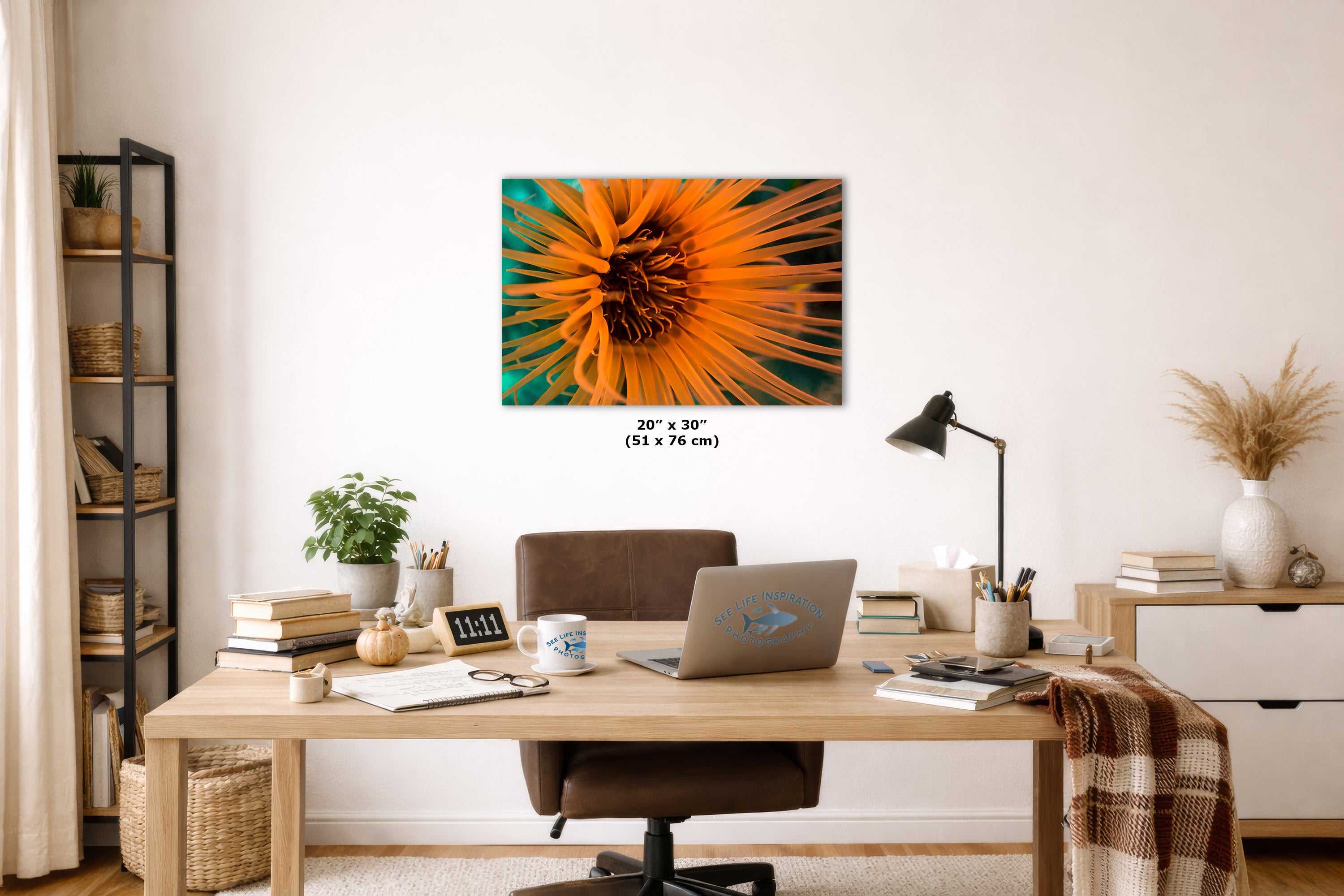 A wall art print featuring a vibrant orange underwater anemone with green and teal accents, displayed in a styled home office setting above a wooden desk with a laptop and chair.