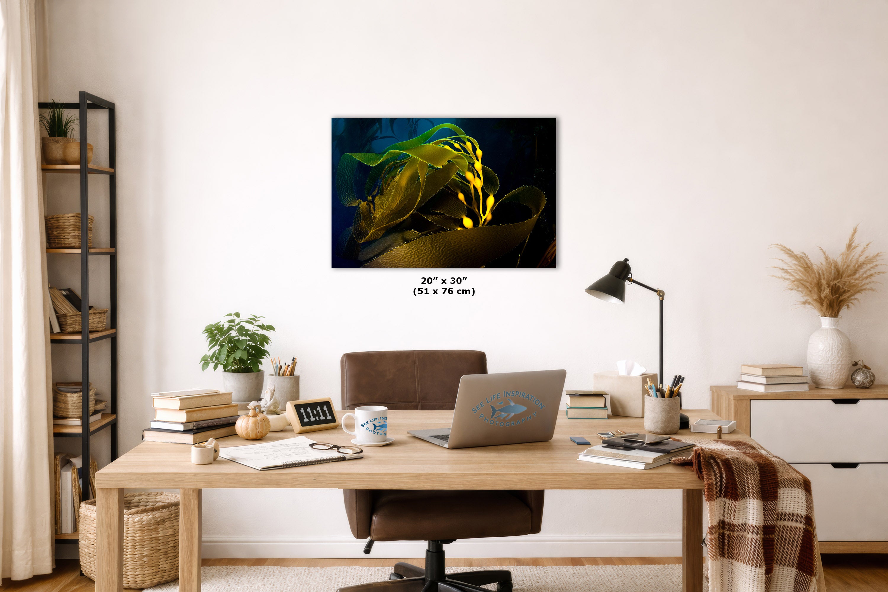 A wall art print showing a close-up of a green leaf with glowing yellow highlights on a dark background, displayed above a wooden desk in a styled home office setting. The print includes size text reading 20 x 30" (51 x 76 cm).