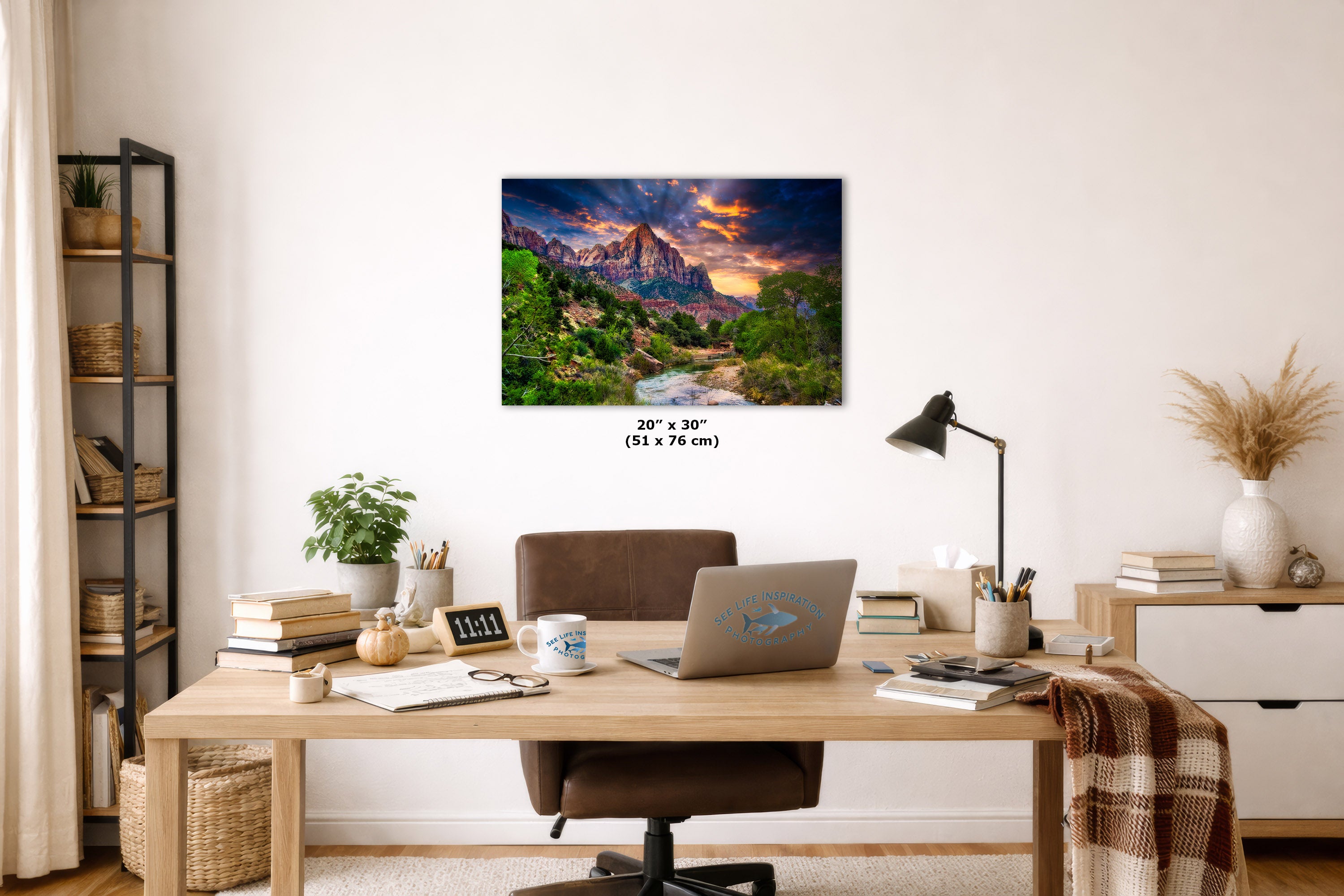 A wall art print showing a colorful mountain landscape with a river and dramatic sunset sky, displayed above a wooden desk in a styled home office. The print includes size text reading 20 x 30 inches.
