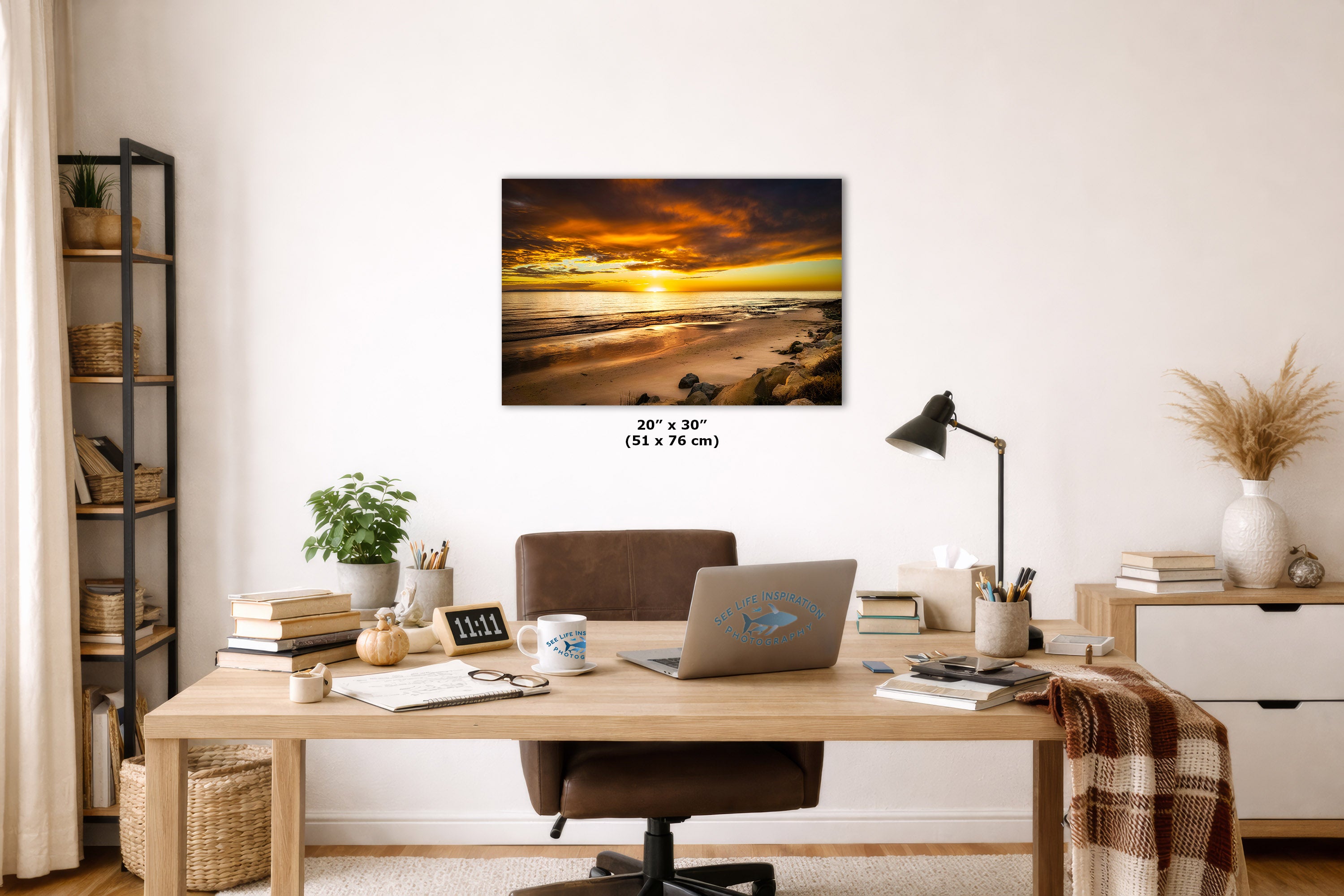 A wall art print showing a dramatic sunset over a beach, displayed above a wooden desk in a styled home office. The print has a black border and includes size text reading 20 x 30 inches (51 x 76 cm) below it.