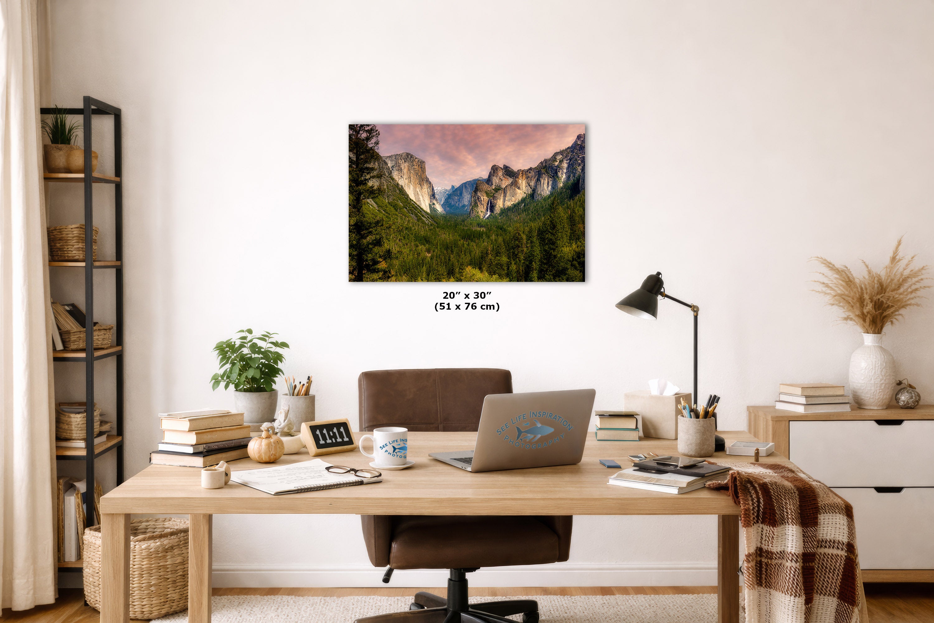 A wall art print depicting a mountain valley landscape with green trees and a pink sky, displayed above a wooden desk in a styled home office. The print includes size text reading 20 x 30 inches (51 x 76 cm).