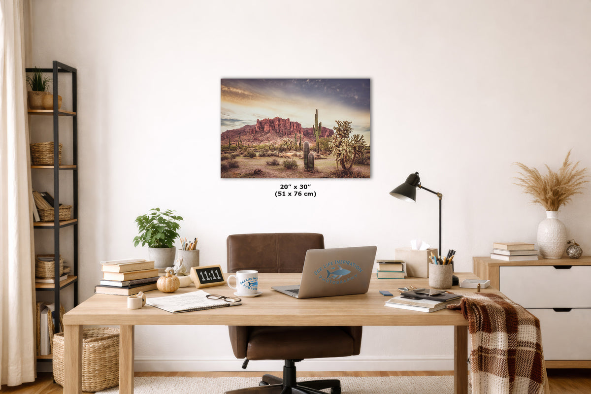 A wall art print depicting a desert canyon landscape with a cactus and sunset sky, displayed above a wooden desk in a styled home office. The print includes size text reading 20 x 30 inches.