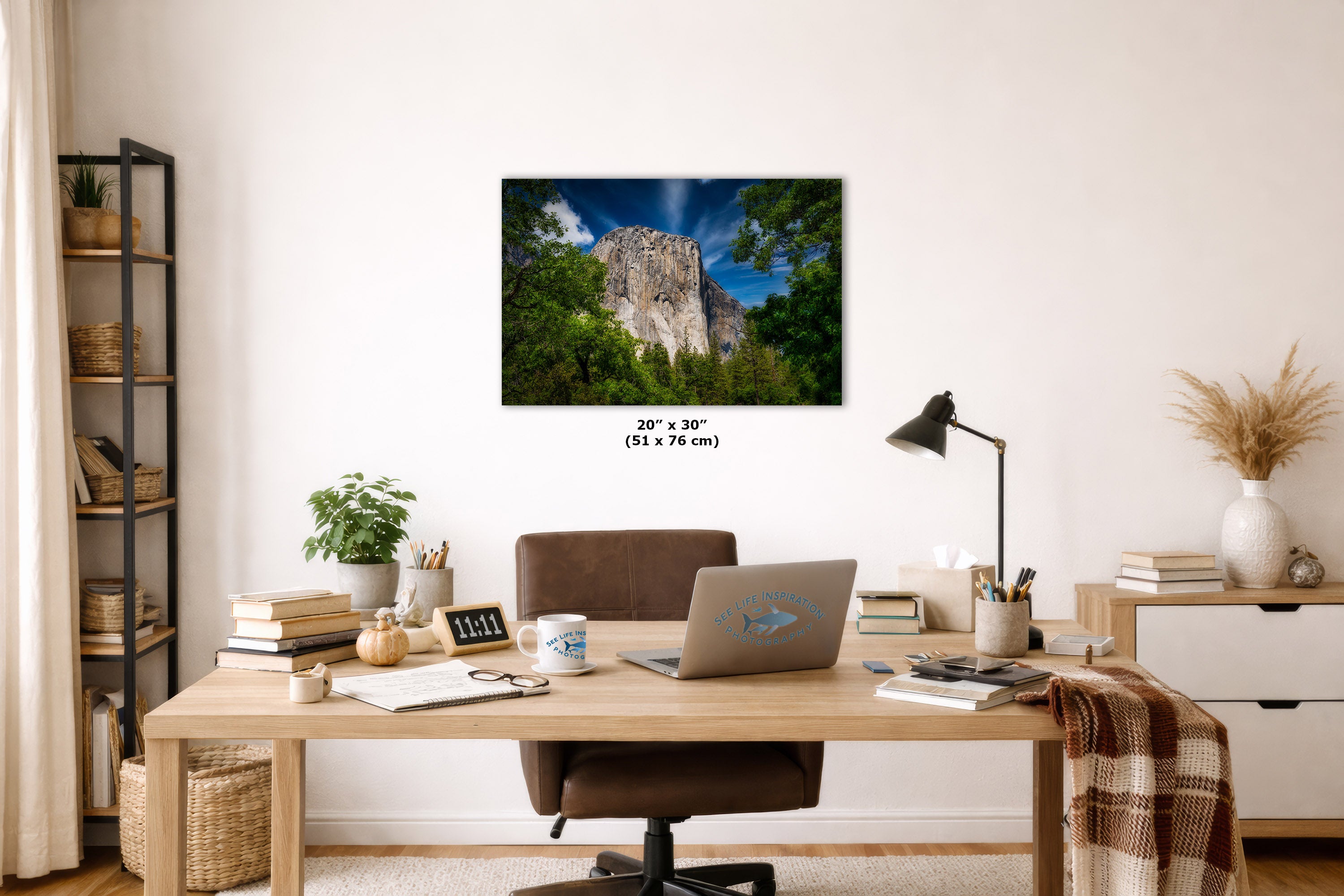 A wall art print showing a rocky cliff surrounded by green trees under a blue sky, displayed above a wooden desk in a styled home office. The print includes size text reading 20 x 30 cm.