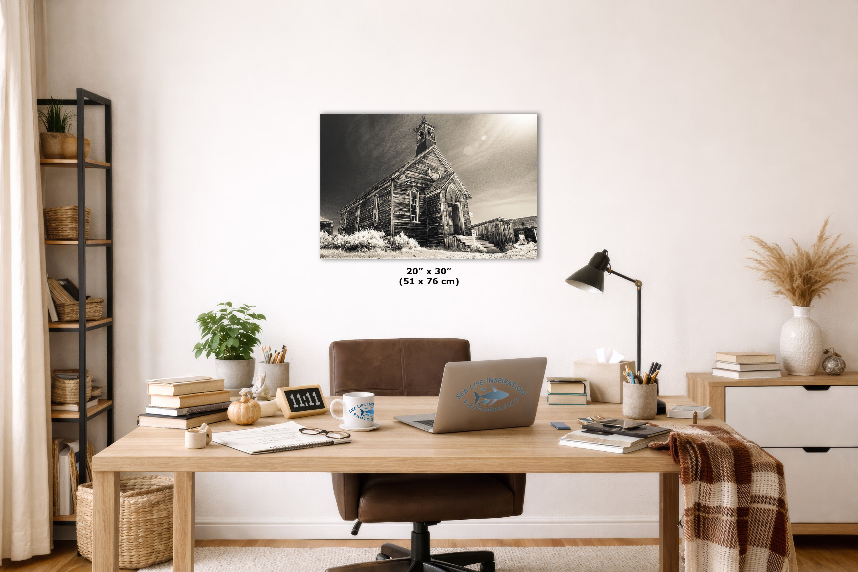 An art print of a rustic old church building in snow, displayed on a white wall in a styled home office setting above a wooden desk with a laptop and chair.