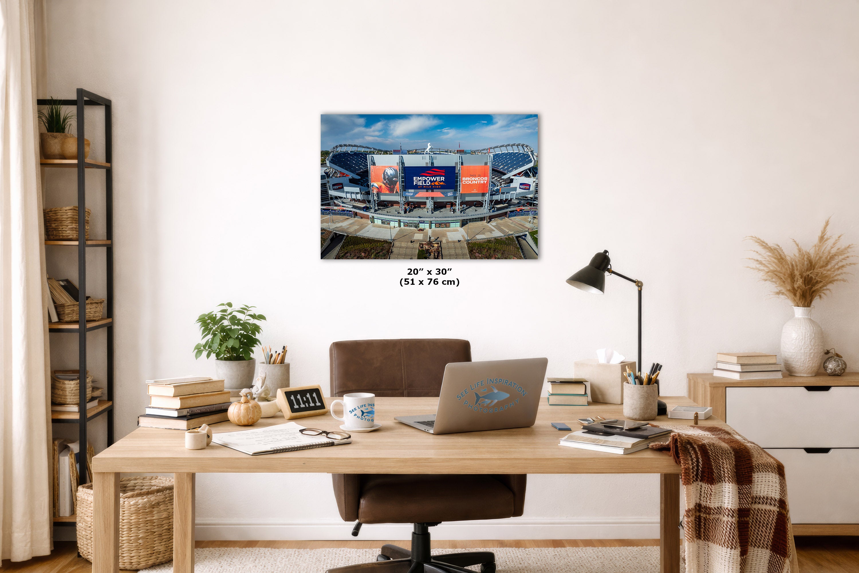 A Broncos Empower Field wall art print depicting the Mile High football stadium under a blue sky, displayed on a white wall above a wooden desk in a styled home office setting. The print includes size text indicating 20 x 30 inches.
