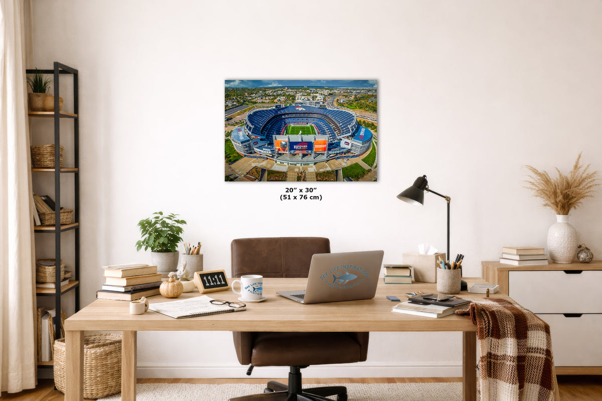A framed wall art print showing an aerial view of a blue stadium, displayed above a wooden desk in a styled home office. The print includes size text reading 20 x 30 inches.