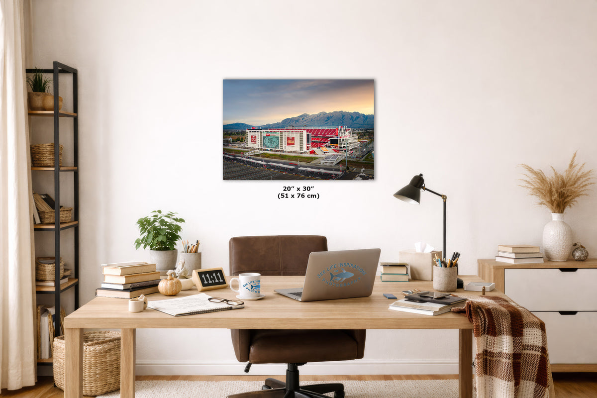 A wall art print featuring a colorful Levi's stadium with mountains in the background, displayed above a wooden desk in a styled home office. The print includes size text reading 20 x 30 cm.