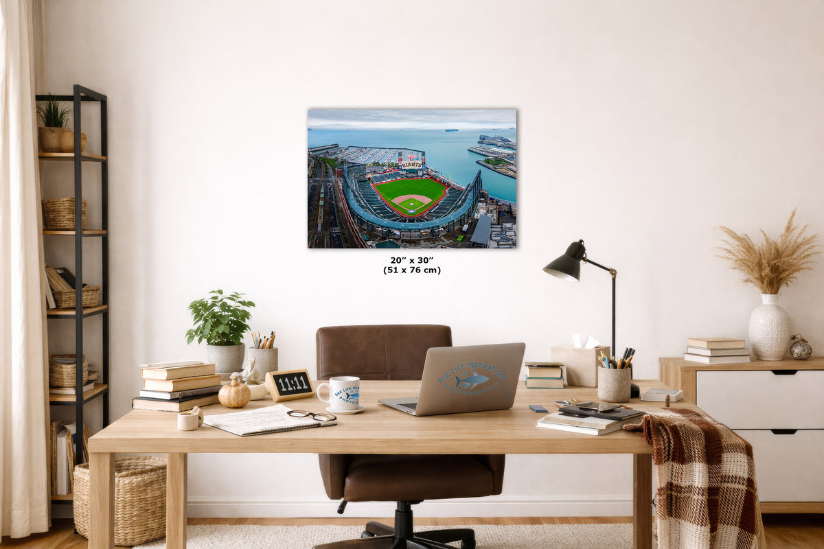 A wall art print showing an aerial view of a baseball stadium and San Francisco Bay, displayed above a wooden desk in a styled home office. The print includes size text reading 20" x 30" (51 x 76 cm) below it.