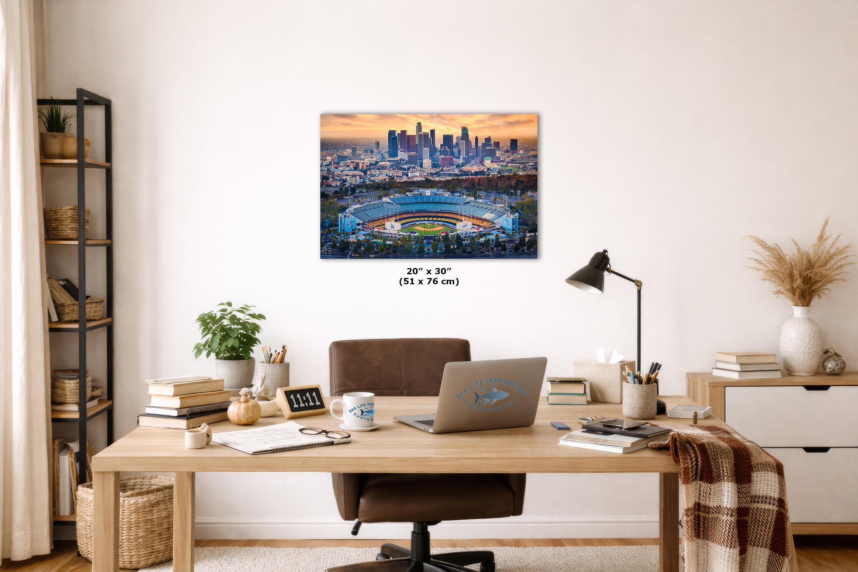 A rectangular Dodger Stadium wall art print showing a city skyline and an illuminated stadium at sunset, displayed above a wooden desk in a styled home office.