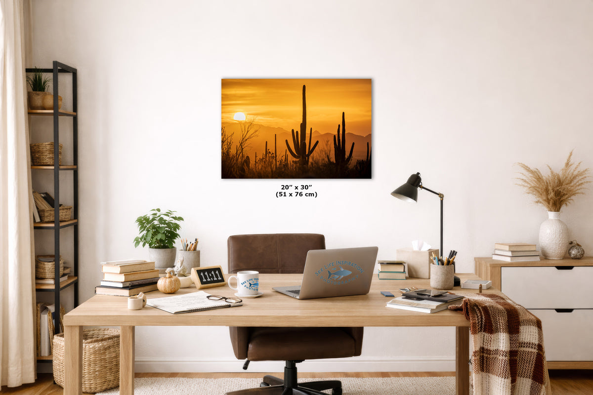 A wall art print showing a desert sunset with tall cacti, displayed above a wooden desk in a styled home office. The print includes size text reading 20 x 30 in (51 x 76 cm) below it.