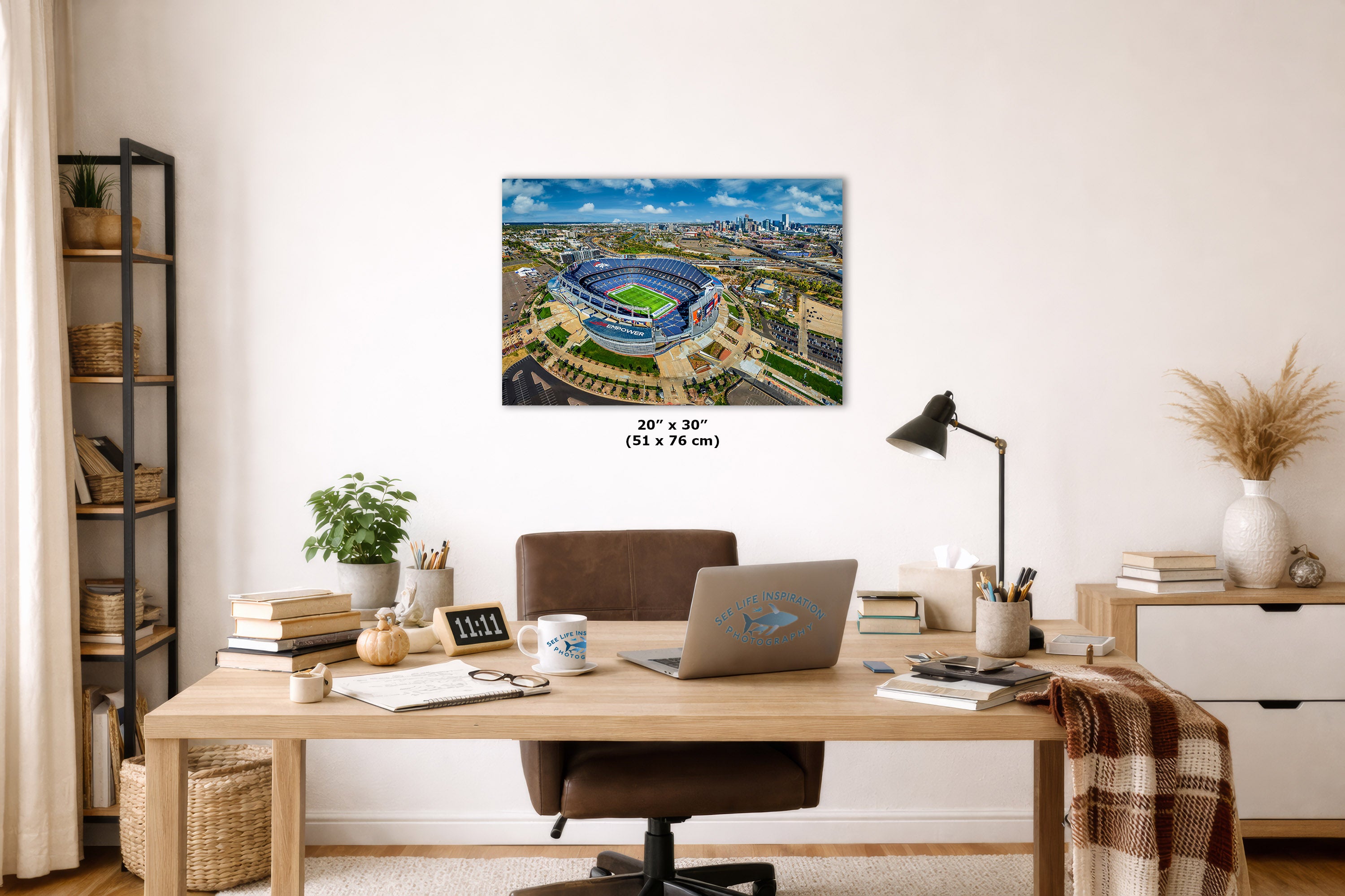 A wall art print showing an aerial view of a blue stadium under a cloudy sky, displayed above a wooden desk in a styled home office. The print includes size text reading 20 x 30 inches (51 x 76 cm).