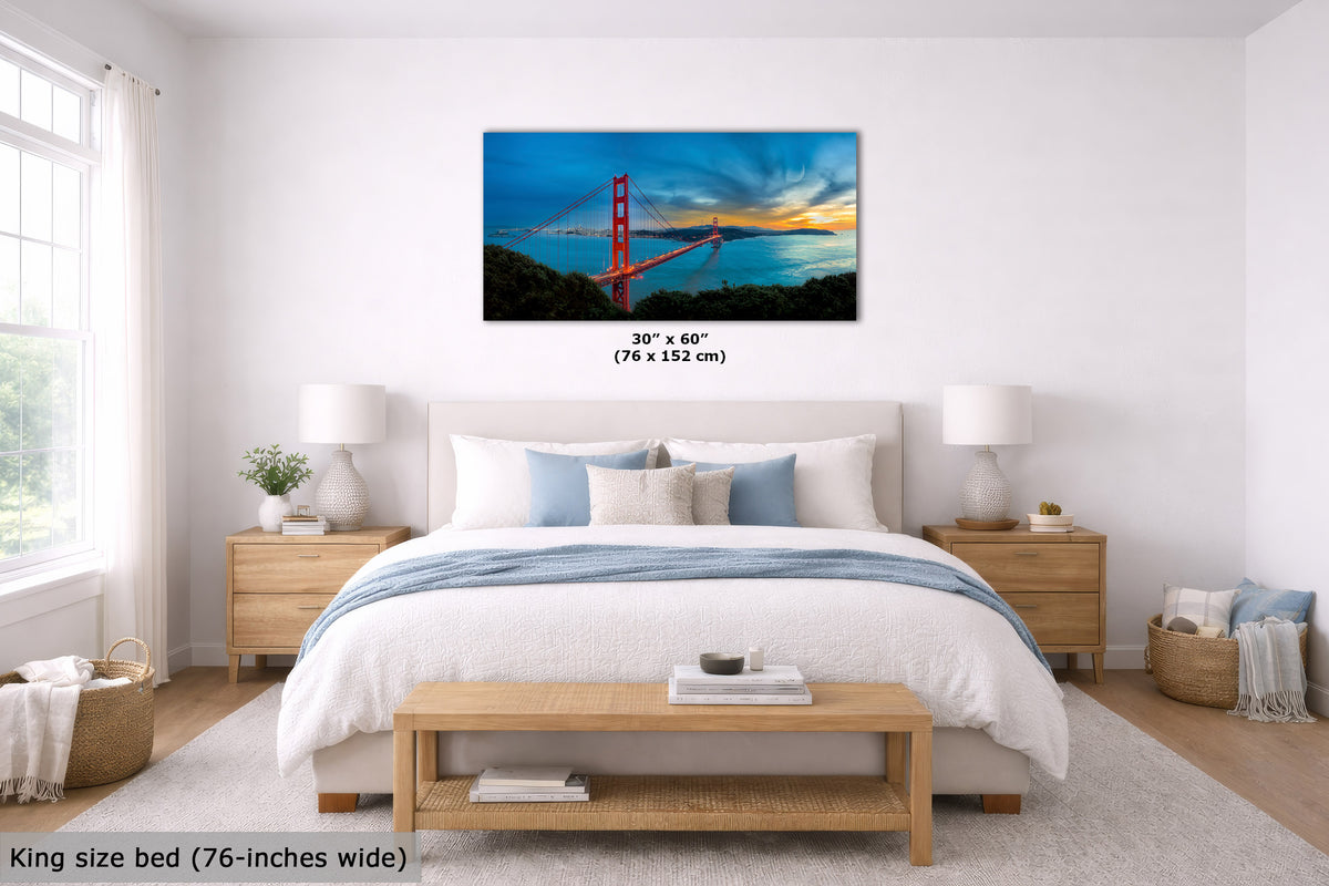 A framed wall art print showing the Golden Gate Bridge over water at sunset, displayed above a neatly made bed in a bright, modern bedroom. The print includes size text reading 30" x 60" (76 x 152 cm), and the room scene includes a bed, nightstands, lamps, and a bench.