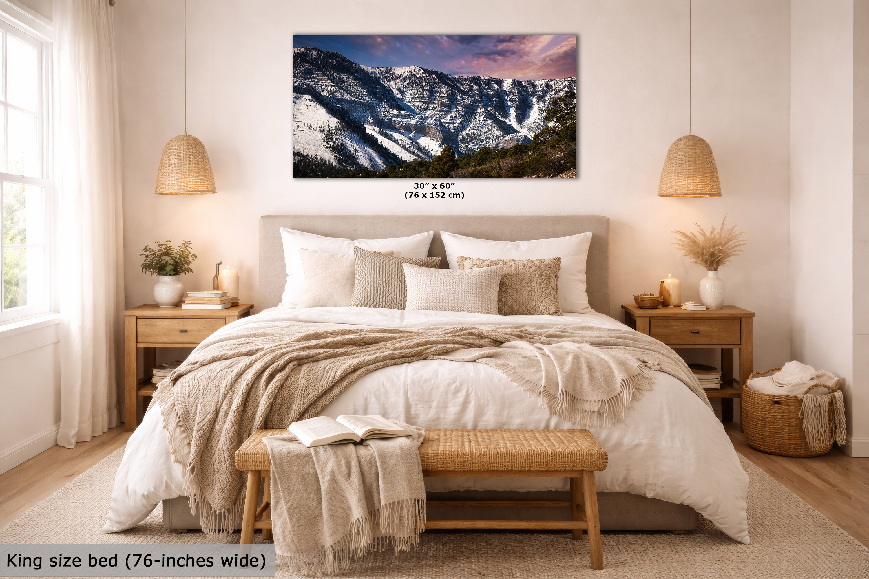 A mountain landscape wall art print displayed above a neatly made bed in a styled bedroom. The print shows snow-covered mountains under a pink and purple sky, with size text below it reading 30 x 60 inches (76 x 152 cm).