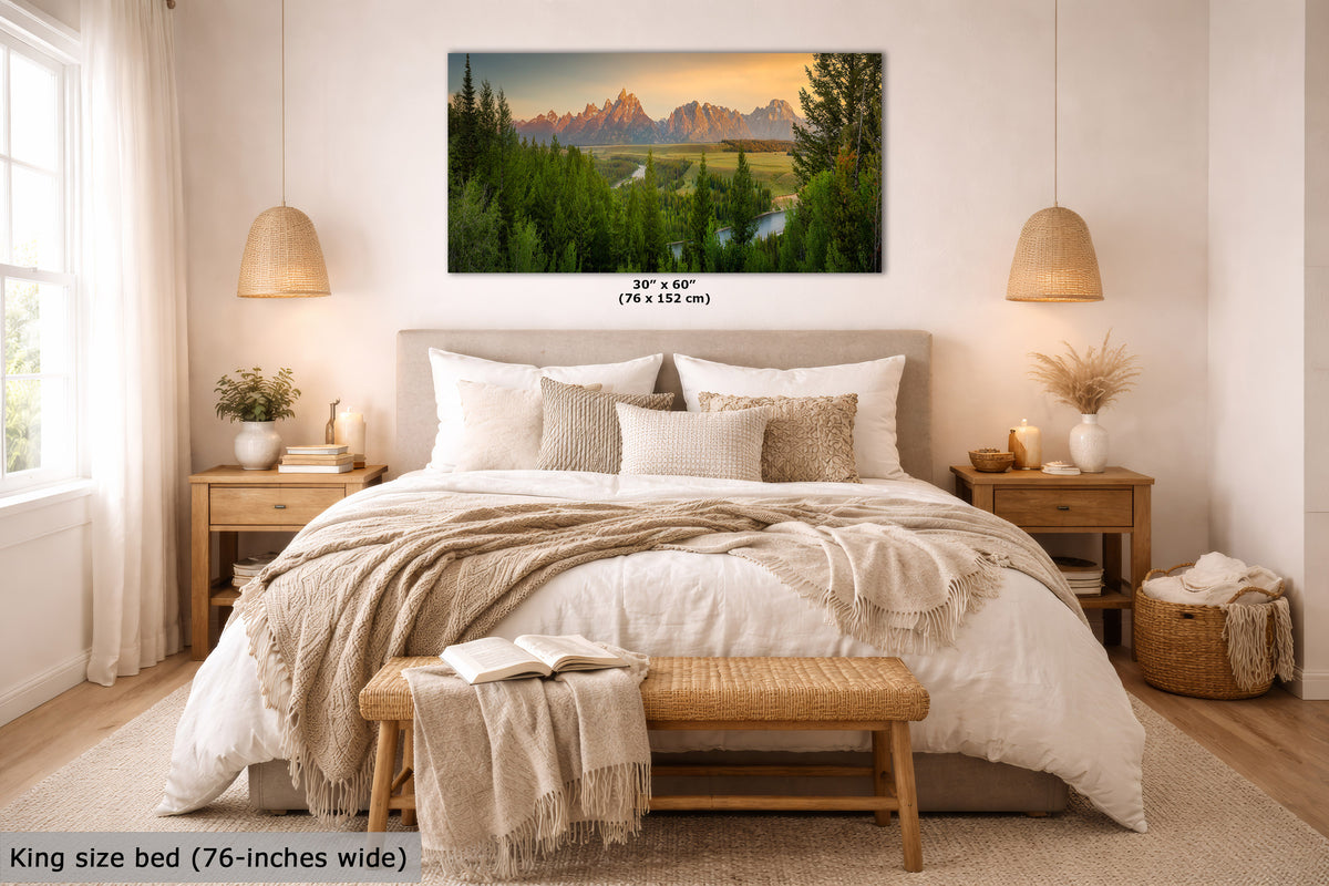 A framed mountain landscape wall art print hangs above a neatly made bed in a bright, modern bedroom. The artwork shows green forest, a river, and jagged mountain peaks at sunset.