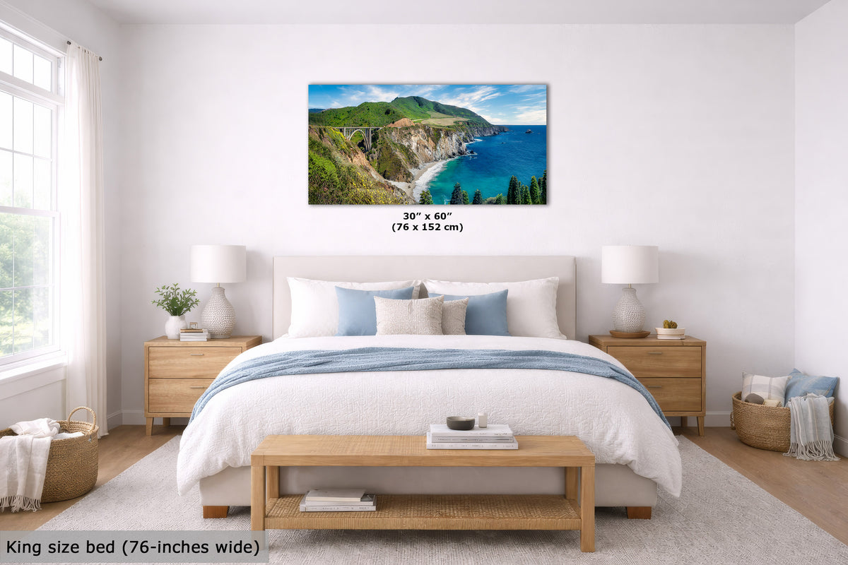A coastal landscape wall art print hangs above a neatly made bed in a bright, modern bedroom. The artwork shows green cliffs and blue ocean water, with size text below it reading 30" x 60" (76 x 152 cm).