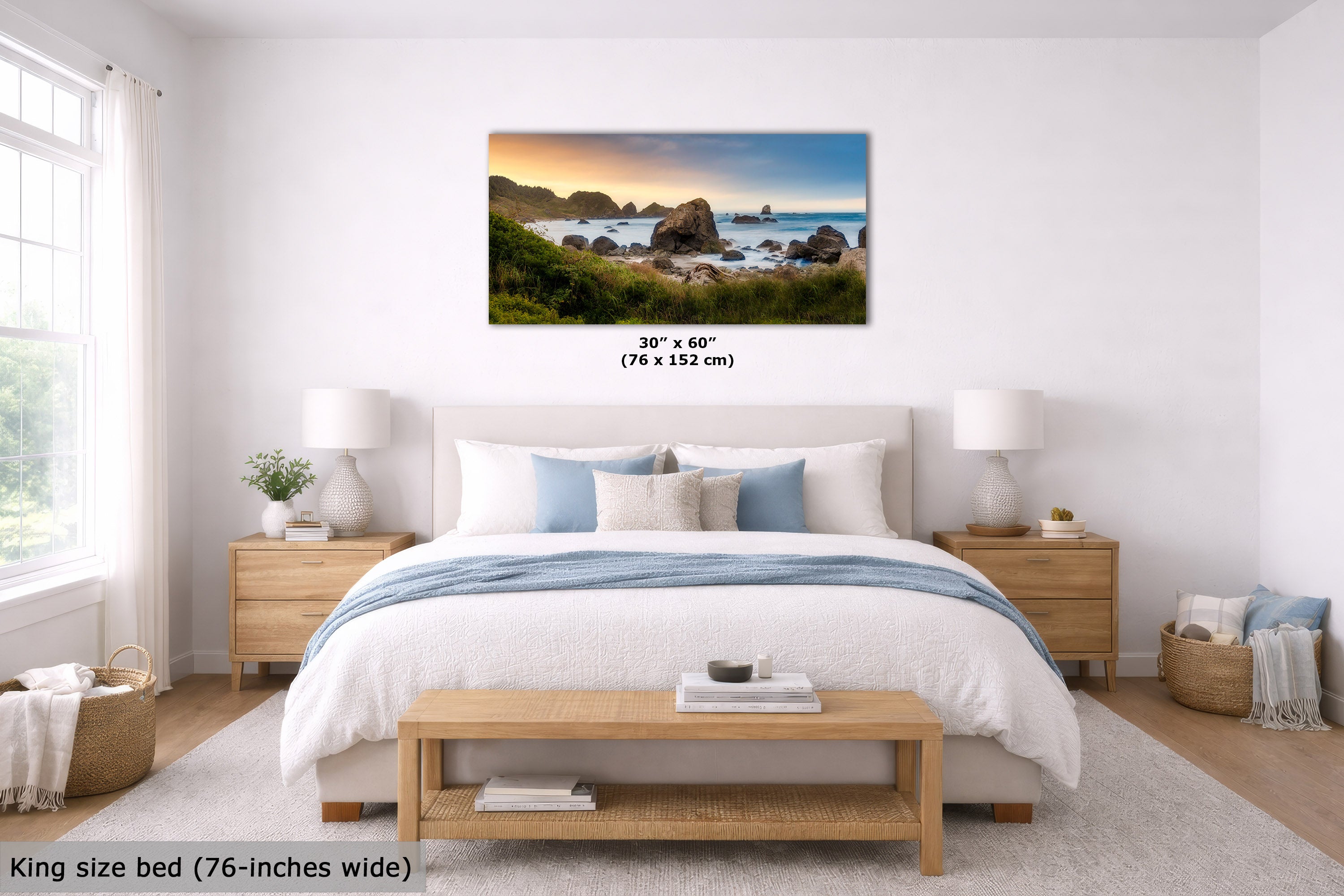 A coastal landscape wall art print hangs above a neatly made bed in a bright, modern bedroom. The print shows a rocky beach at sunset with blue water and green cliffs.