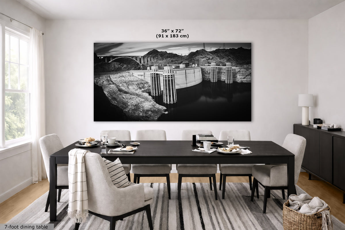 A large black and white wall art print depicting a dam and bridge landscape, displayed in a modern dining room setting above a dark wood dining table and chairs. The print includes a size overlay indicating 36 x 72 inches (91 x 183 cm).