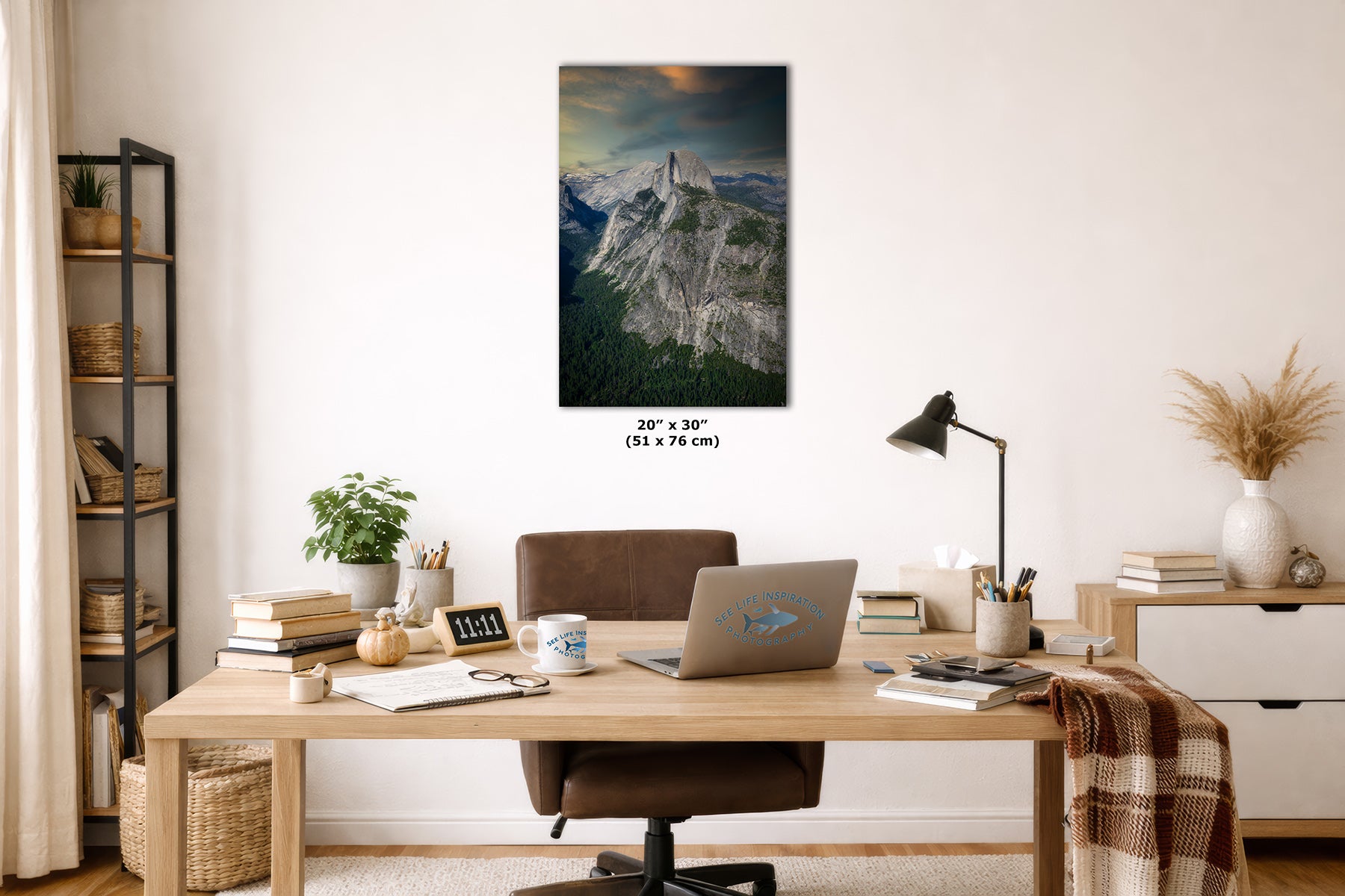 A vertical wall art print of Half Dome in Yosemite, depicting a dramatic mountain landscape with a glowing sunset, displayed on a white wall above a wooden desk in a modern home office setting.