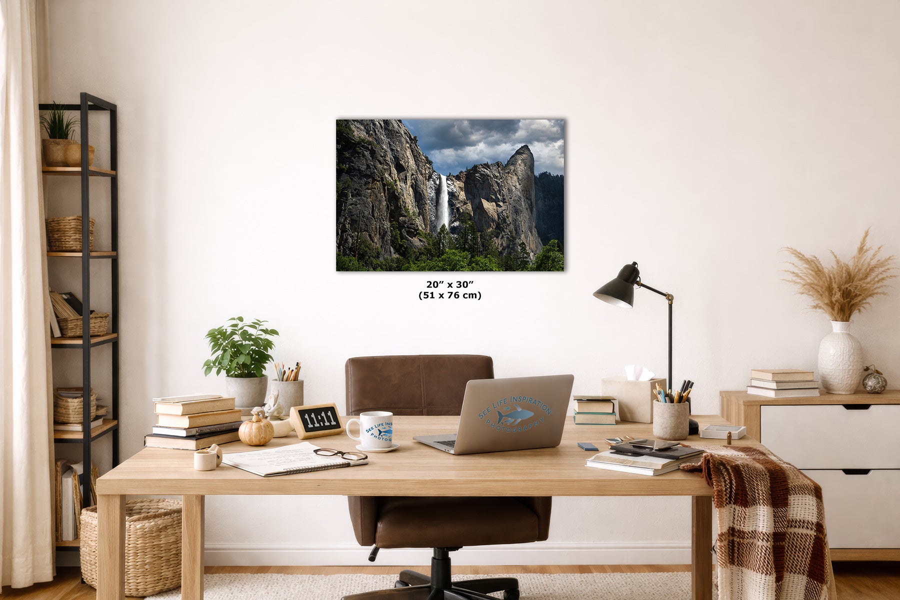A framed wall art print showing Yosemite's Bridalveil Waterfall between rocky cliffs hangs above a wooden desk in a styled home office. The print includes size text reading 24 x 30 inches (51 x 76 cm).