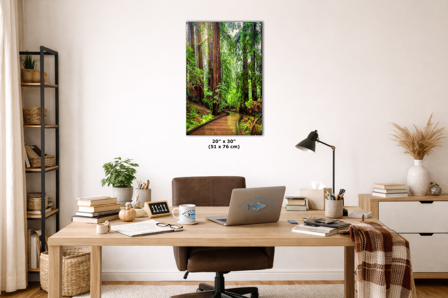 A vertical canvas wall art print depicting a lush green Redwood forest path, styled in a modern home office setting above a wooden desk with a laptop, lamp, and other office accessories.