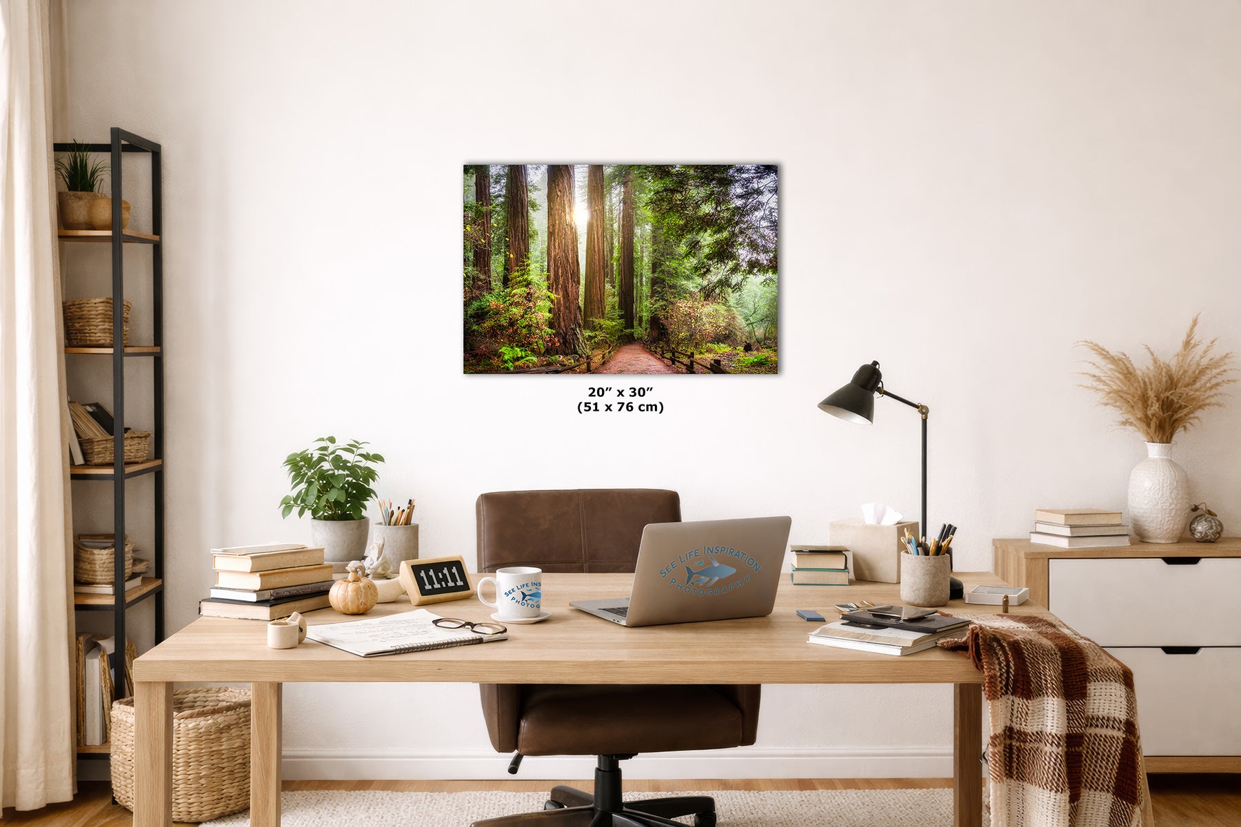 A framed wall art print depicting a sunlit California Redwood forest path hangs above a wooden desk in a styled home office. The print is shown as the product, with room decor including a chair, laptop, lamp, books, and shelving visible around it.