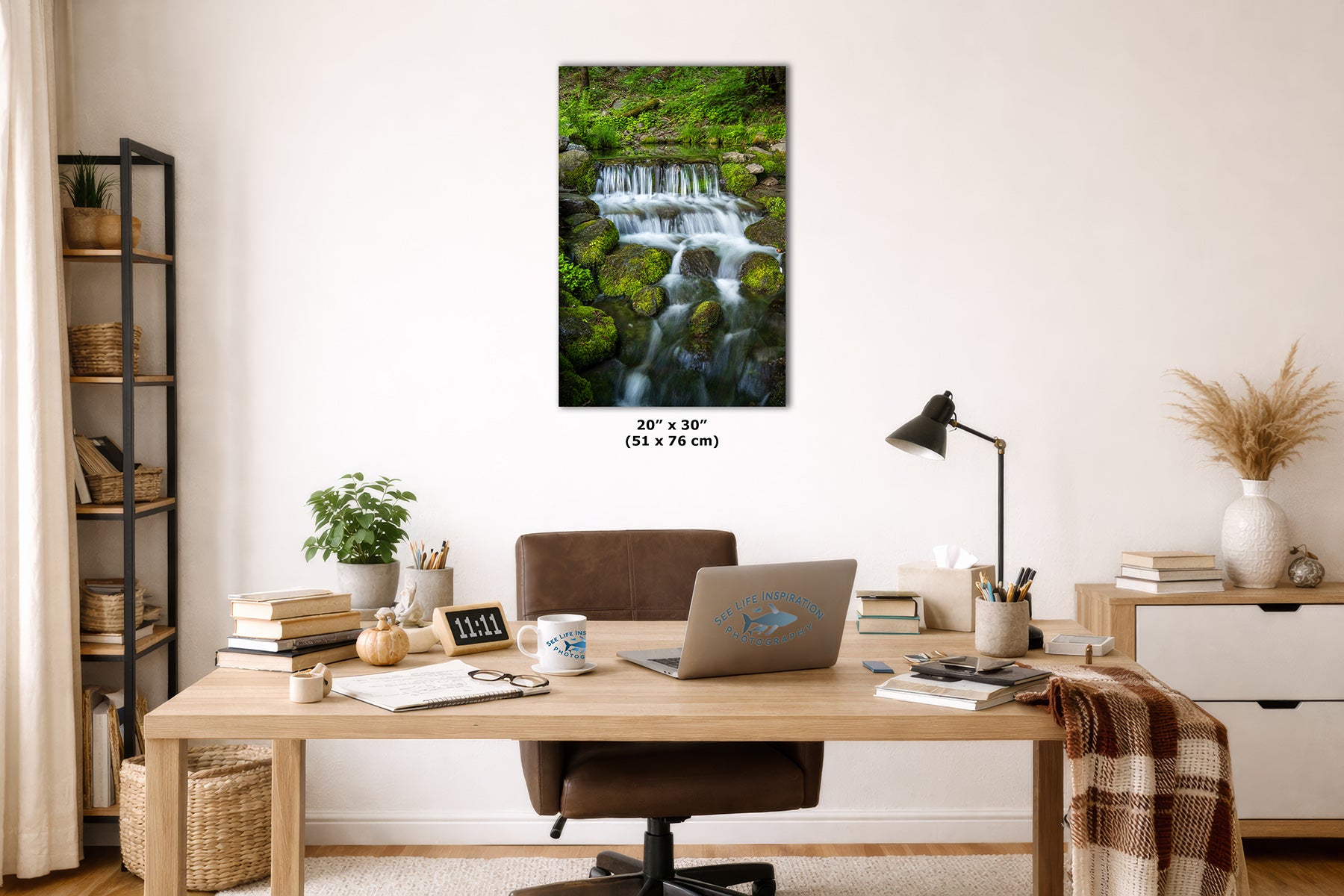 A wall art print depicting a lush green waterfall cascading over mossy rocks, displayed in a styled home office setting above a wooden desk with a laptop, lamp, and other office accessories.
