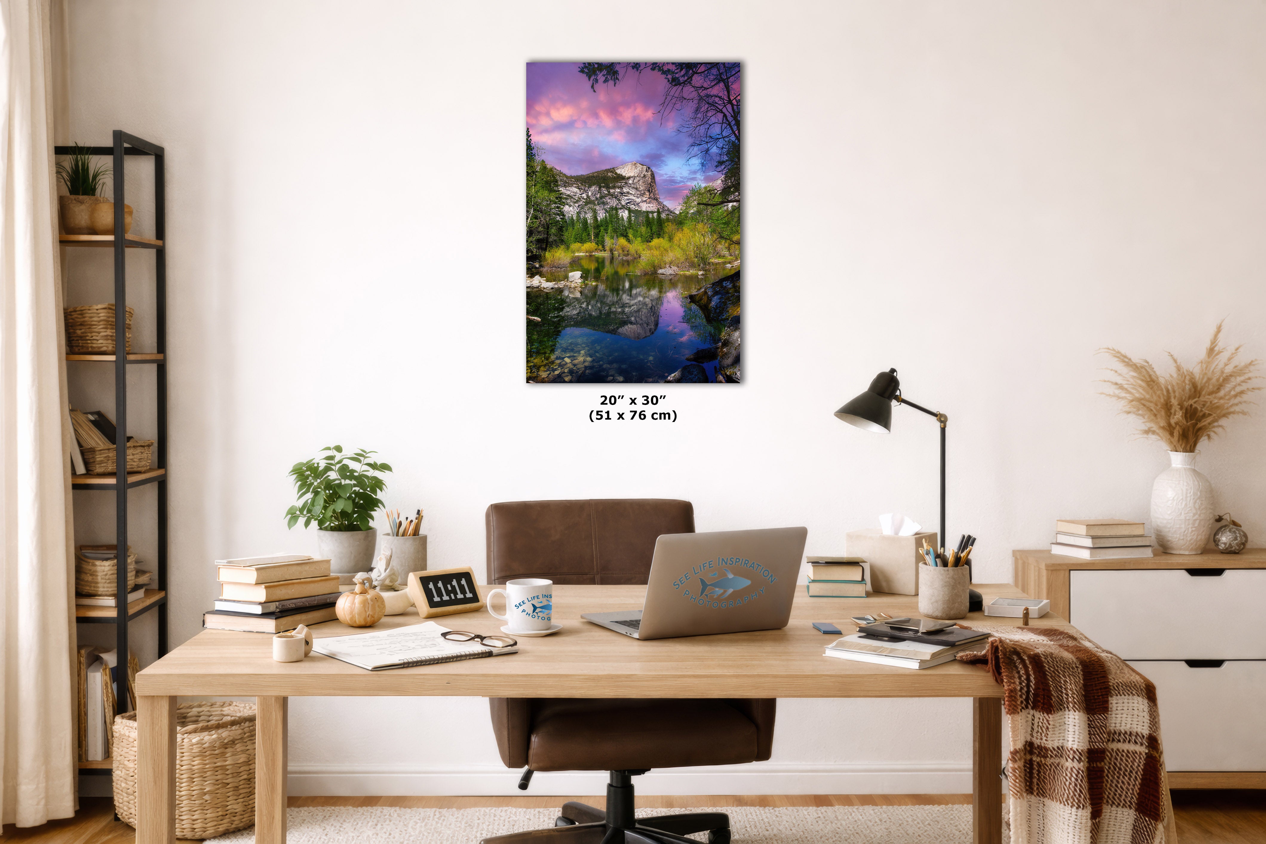 A framed vertical canvas print of a colorful landscape with a lake, trees, and a pink-purple sky, displayed on a white wall above a wooden desk in a styled home office setting.