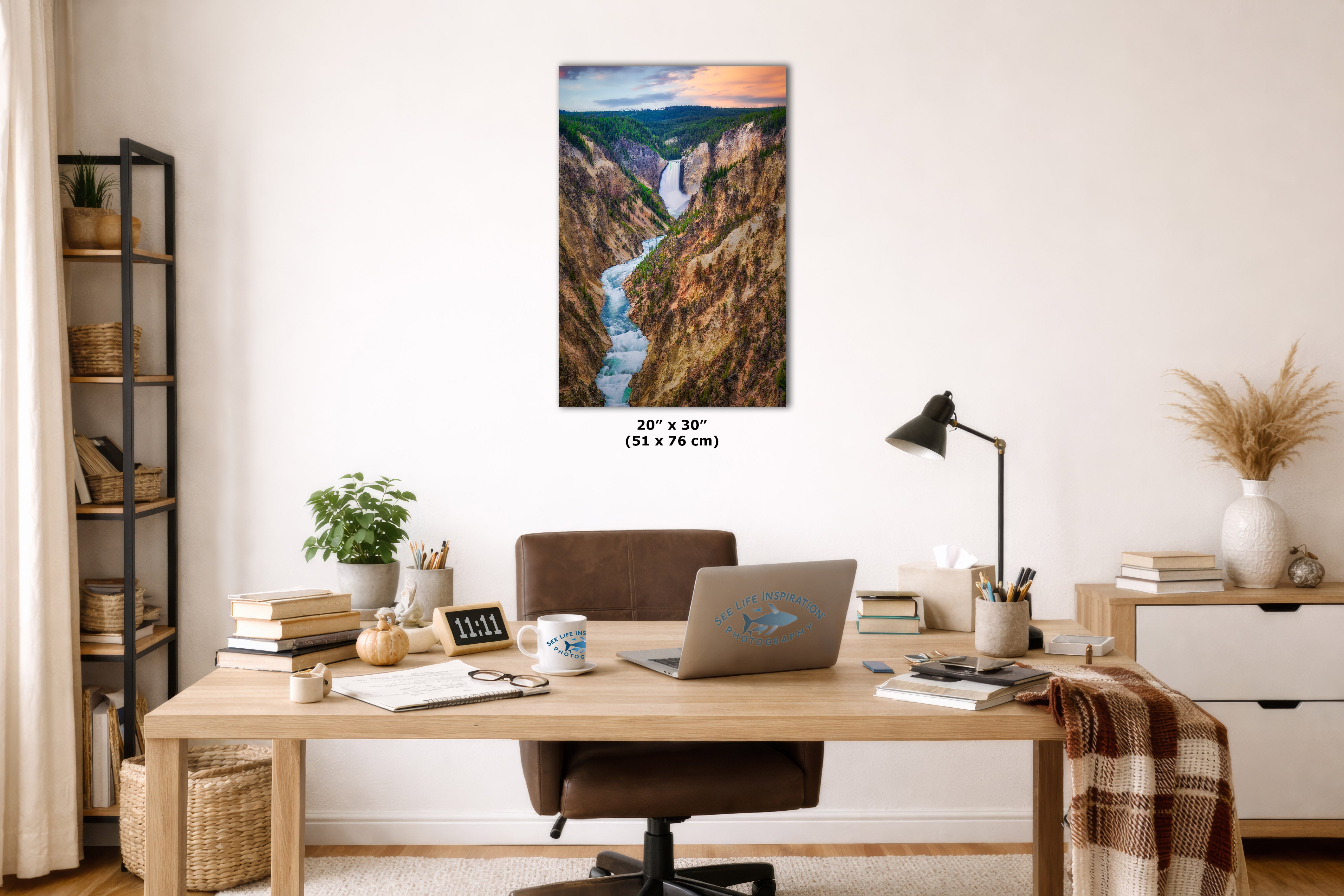 A framed vertical landscape art print depicting a waterfall in a canyon, displayed on a white wall above a wooden desk in a styled home office setting.