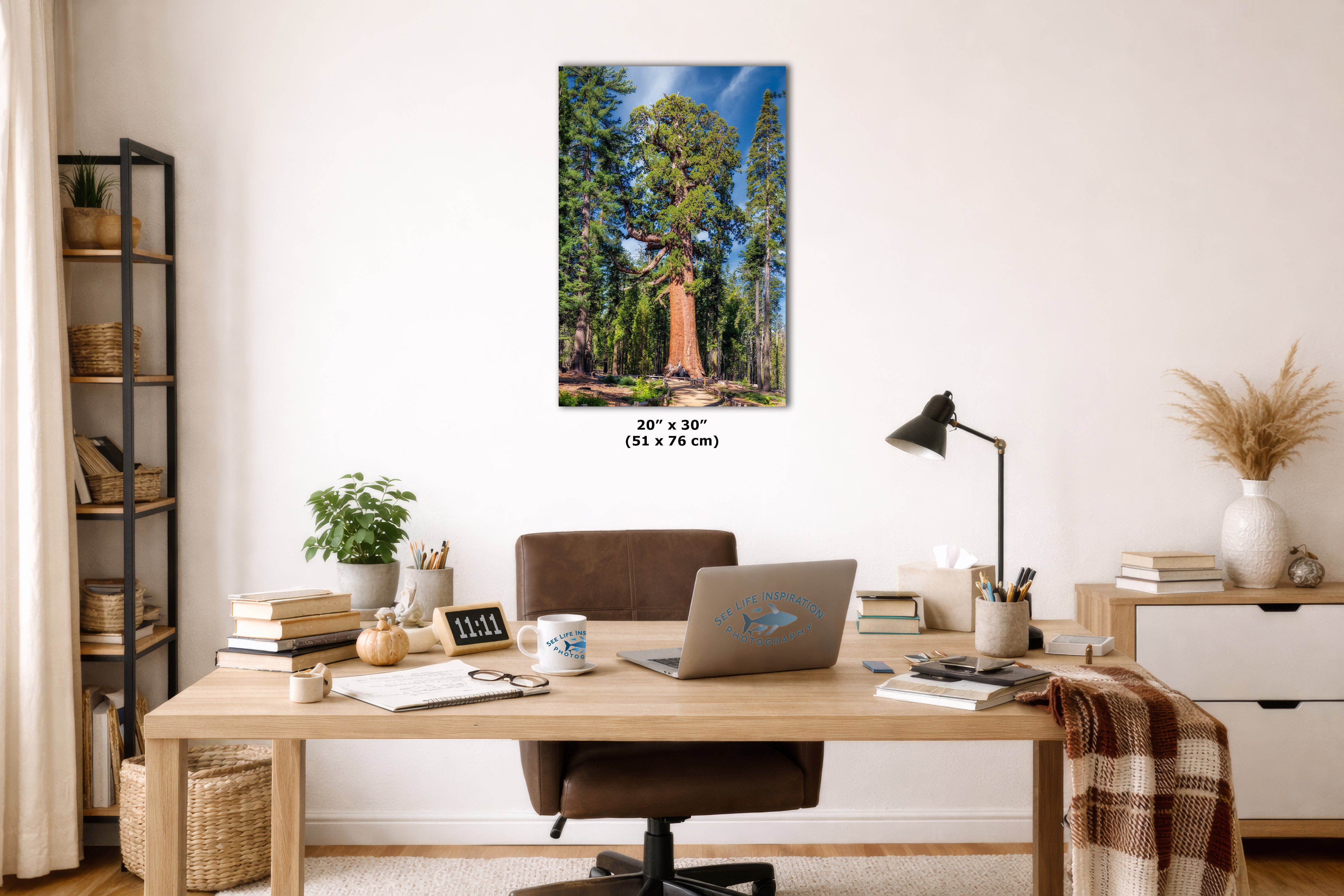 A vertical canvas wall art print depicting a forest scene with a large tree, displayed on a white wall above a wooden desk in a styled home office setting.