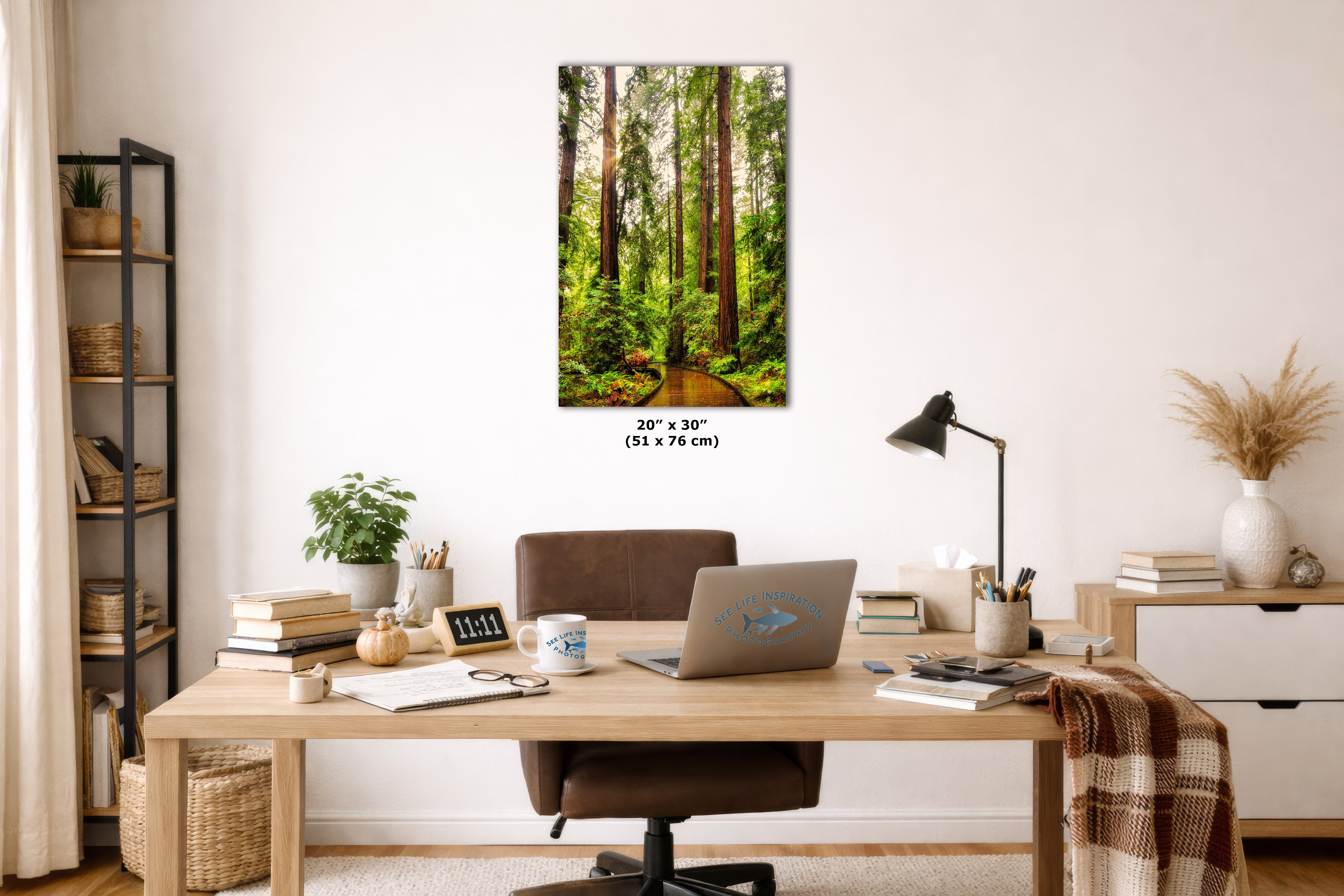A vertical canvas wall art print depicting a lush green forest path, styled in a modern home office setting above a wooden desk with a laptop and chair.