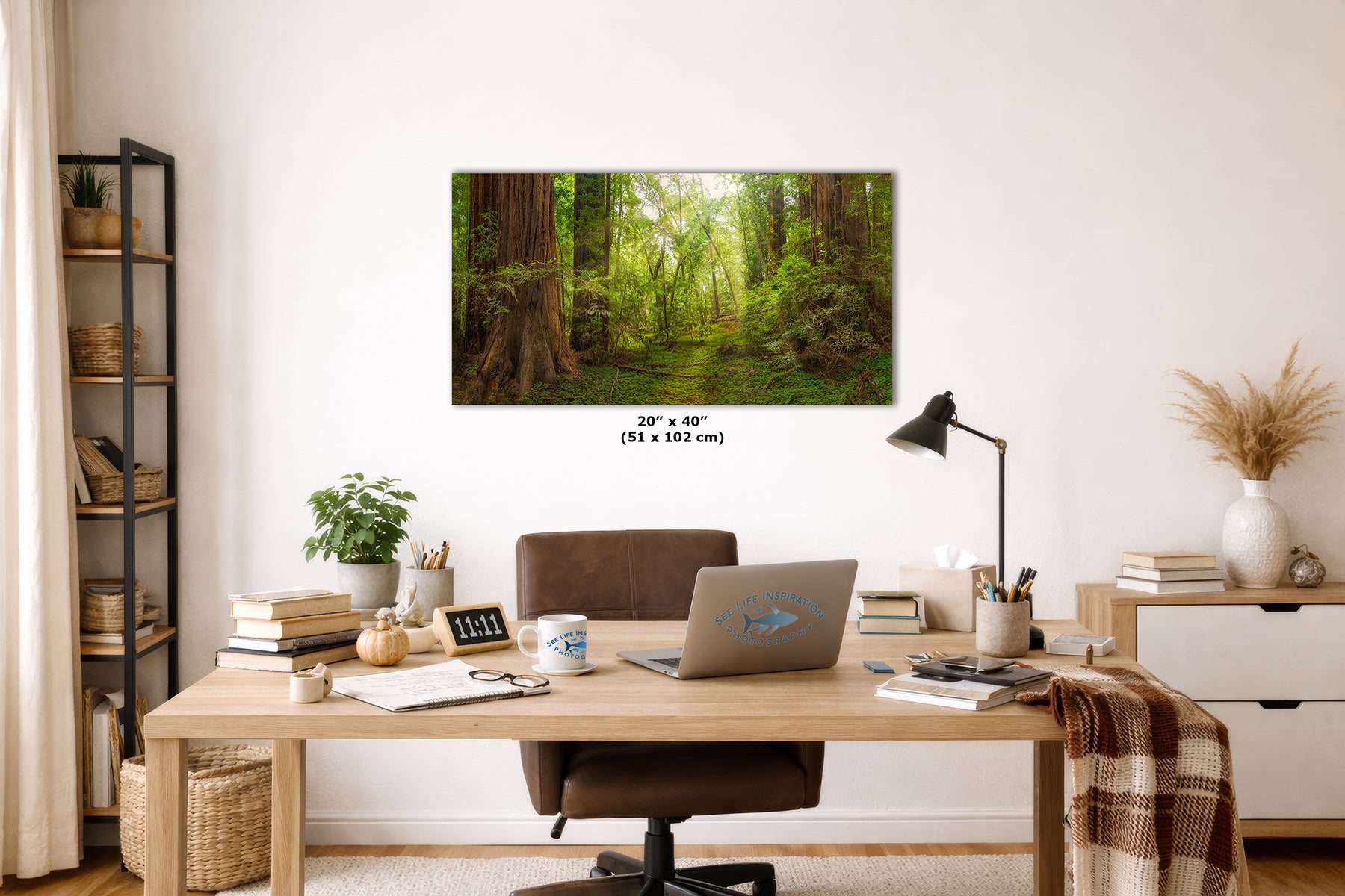 A wall art print depicting a lush green Redwood forest path hangs above a wooden desk in a styled home office. The print is shown in a real interior setting with a chair, laptop, lamp, books, and shelving, and includes size text below the artwork.