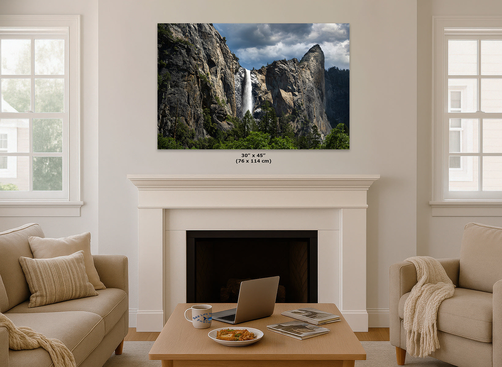 A wall art print of Yosemite's Bridalveil Falls, shown hanging above a white fireplace mantel in a styled living room with beige sofas and a coffee table. The print includes a size label reading 30 x 45 in (70 x 114 cm).
