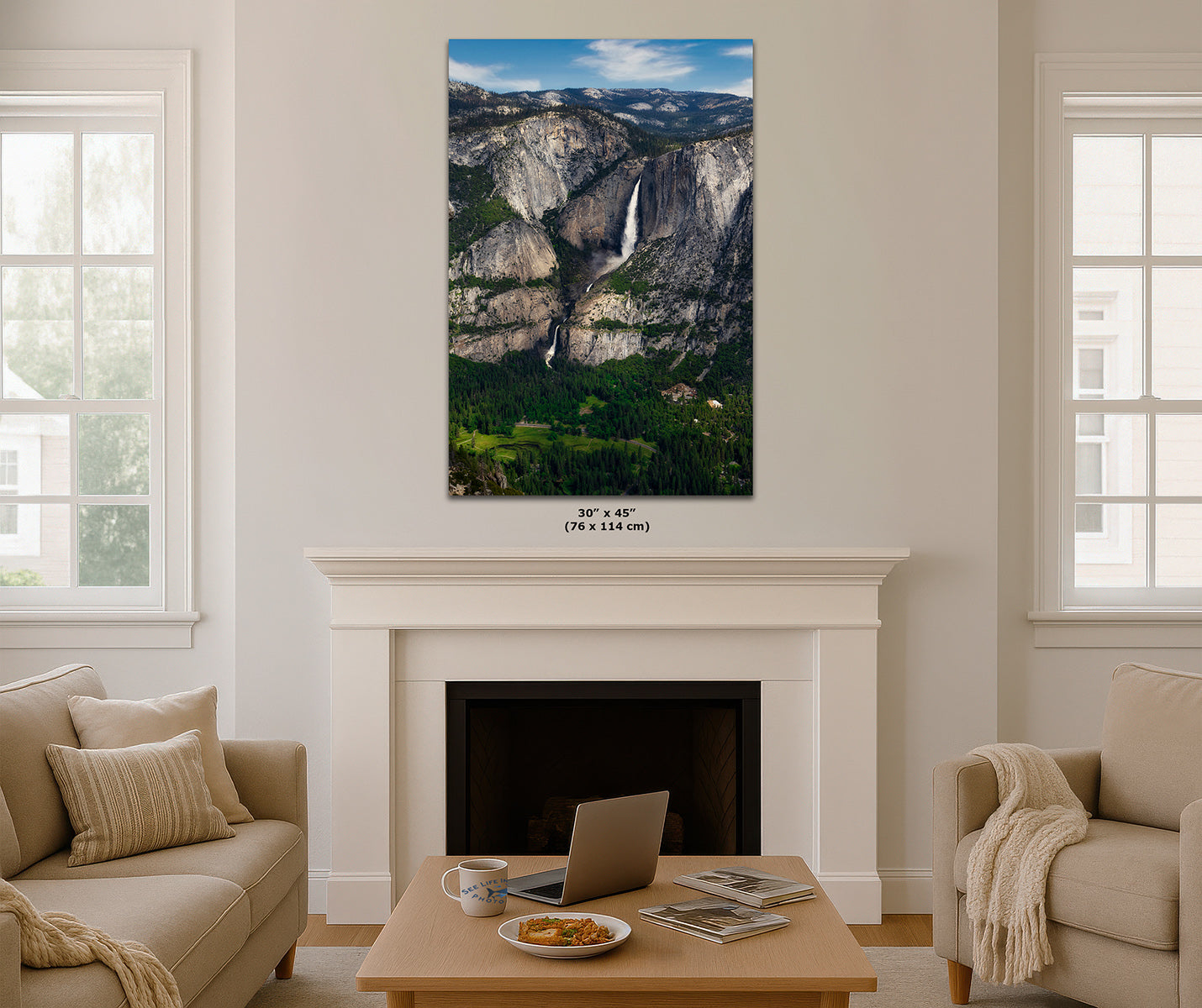 A vertical print of Yosemite Falls landscape, displayed on a white wall above a fireplace in a modern living room setting with beige sofas and a wooden coffee table.