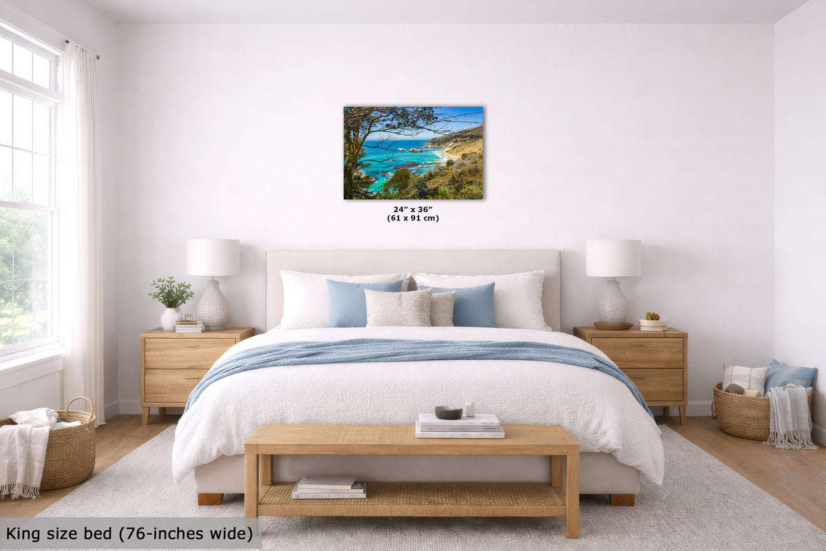 A framed coastal landscape wall art print hangs above a neatly made bed in a bright, modern bedroom. The artwork depicts turquoise ocean water, sandy shorelines, and dark tree branches.