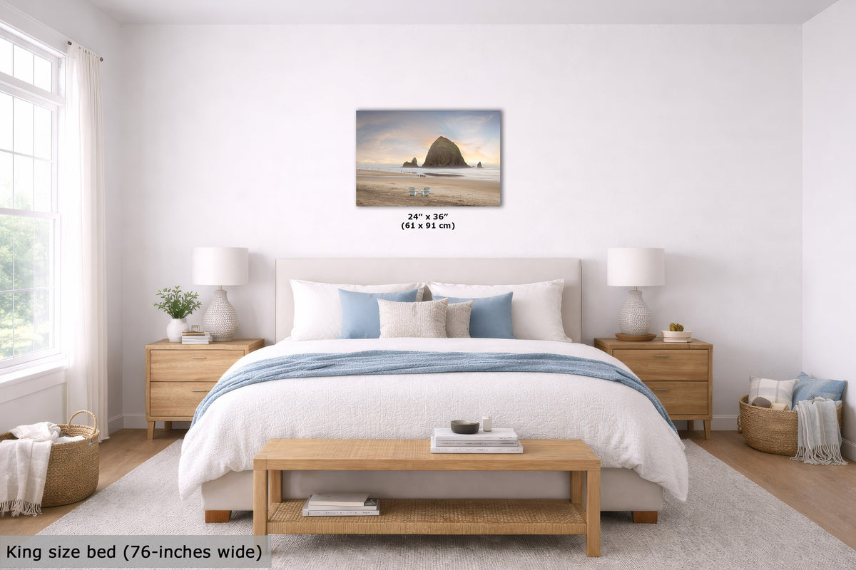 A framed beach landscape wall art print is displayed above a neatly made bed in a bright, modern bedroom. The print shows a coastal rock formation at sunset.