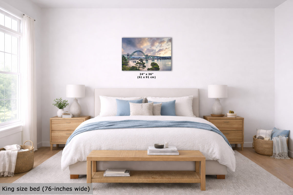 A wall art print depicting a scenic bridge over water under a dramatic cloudy sky, displayed above a neatly made bed in a bright, modern bedroom. The print includes size text reading 24" x 36" (61 x 91 cm).