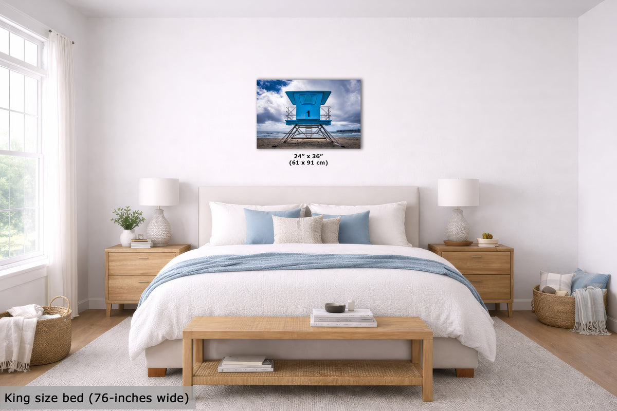 A wall art print showing a bright blue lifeguard tower over water, displayed above a neatly made bed in a bright, modern bedroom. The print includes size text reading 24" x 36" (61 x 91 cm).