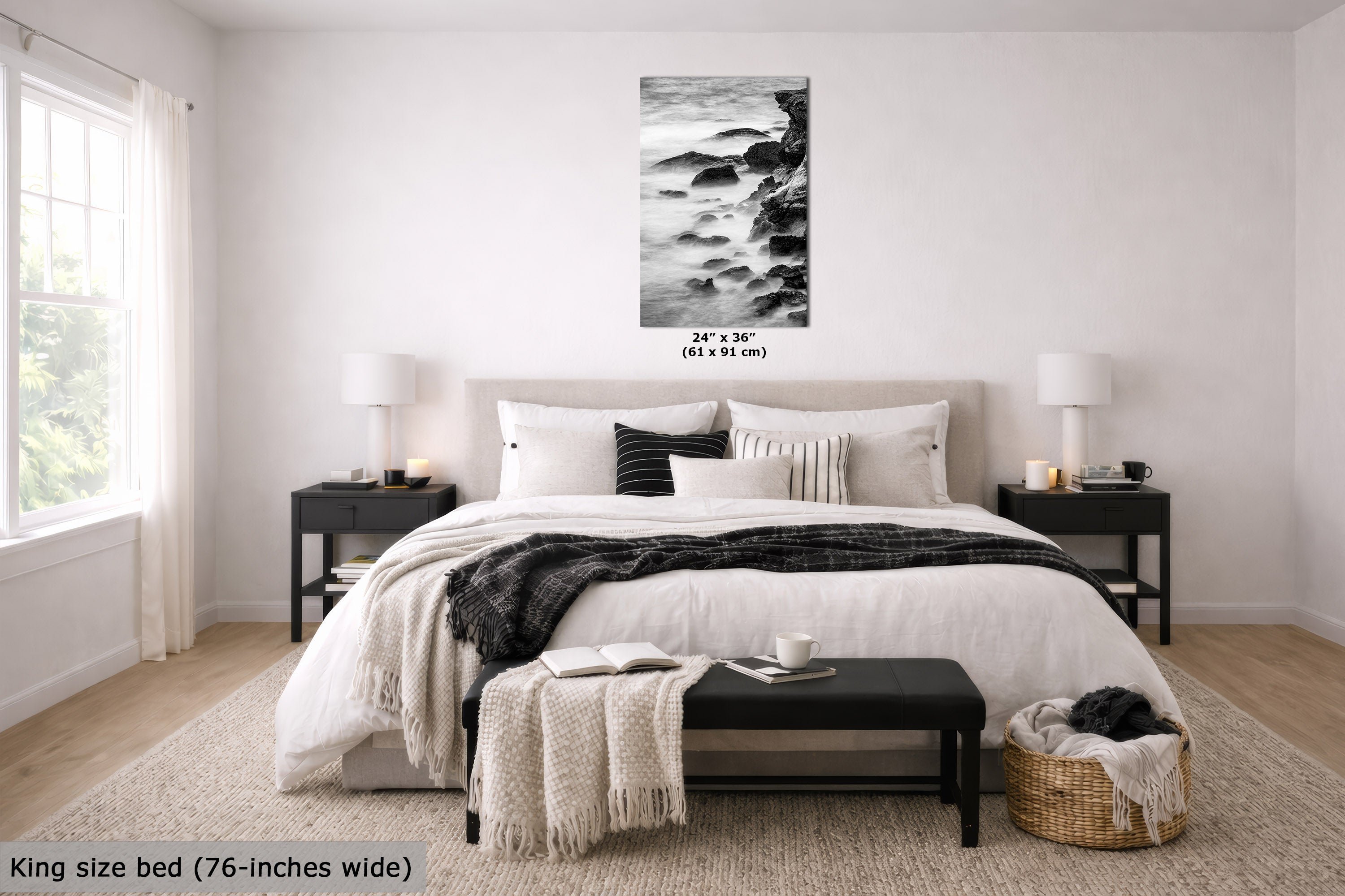 A black and white coastal wave art print displayed on a light gray wall above a neatly made bed in a modern, bright bedroom. The print features a rocky shoreline with waves crashing, and text below it reads '24" x 36" (61 x 91 cm)'.