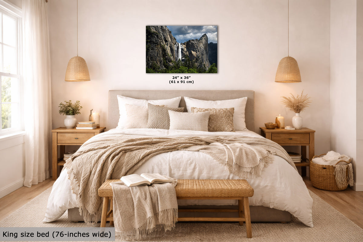 A wall art print showing a rocky mountain landscape with a waterfall, displayed above a neatly made bed in a bright, neutral-toned bedroom. The print includes size text reading 24 x 36 inches (61 x 91 cm), and the room scene also includes a bed, nightstands, lamps, and decorative textiles.