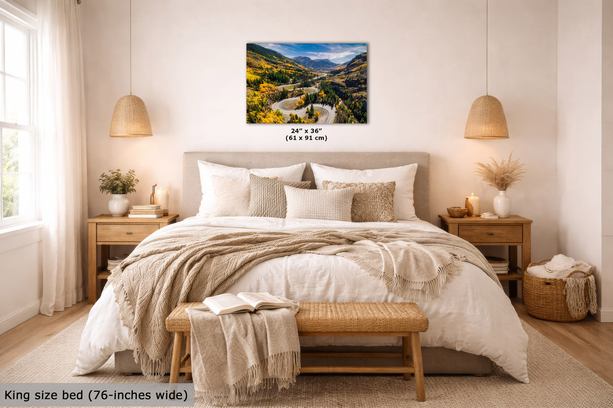 A framed wall art print showing a mountain landscape with a winding road and colorful foliage, displayed above a neatly made bed in a bright, modern bedroom. The print includes size text reading 24 x 36 inches (61 x 91 cm).