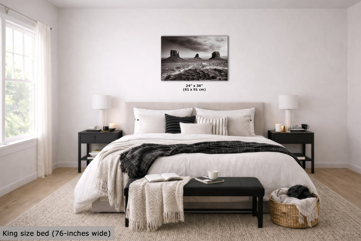 A black and white landscape canvas print of desert rock formations is displayed on a light gray wall above a neatly made bed in a modern, bright bedroom. The text '24" x 36" (61 x 91 cm)' is printed directly below the canvas, and a text overlay in the bottom left corner reads 'King size bed (76-inches wide)'.
