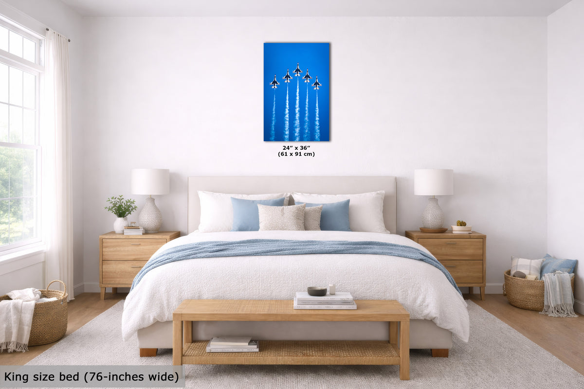 A bedroom scene with a bed, nightstands, lamps, and a blue wall art print centered above the headboard. The print shows five jets flying upward on a bright blue background, with size text below it.