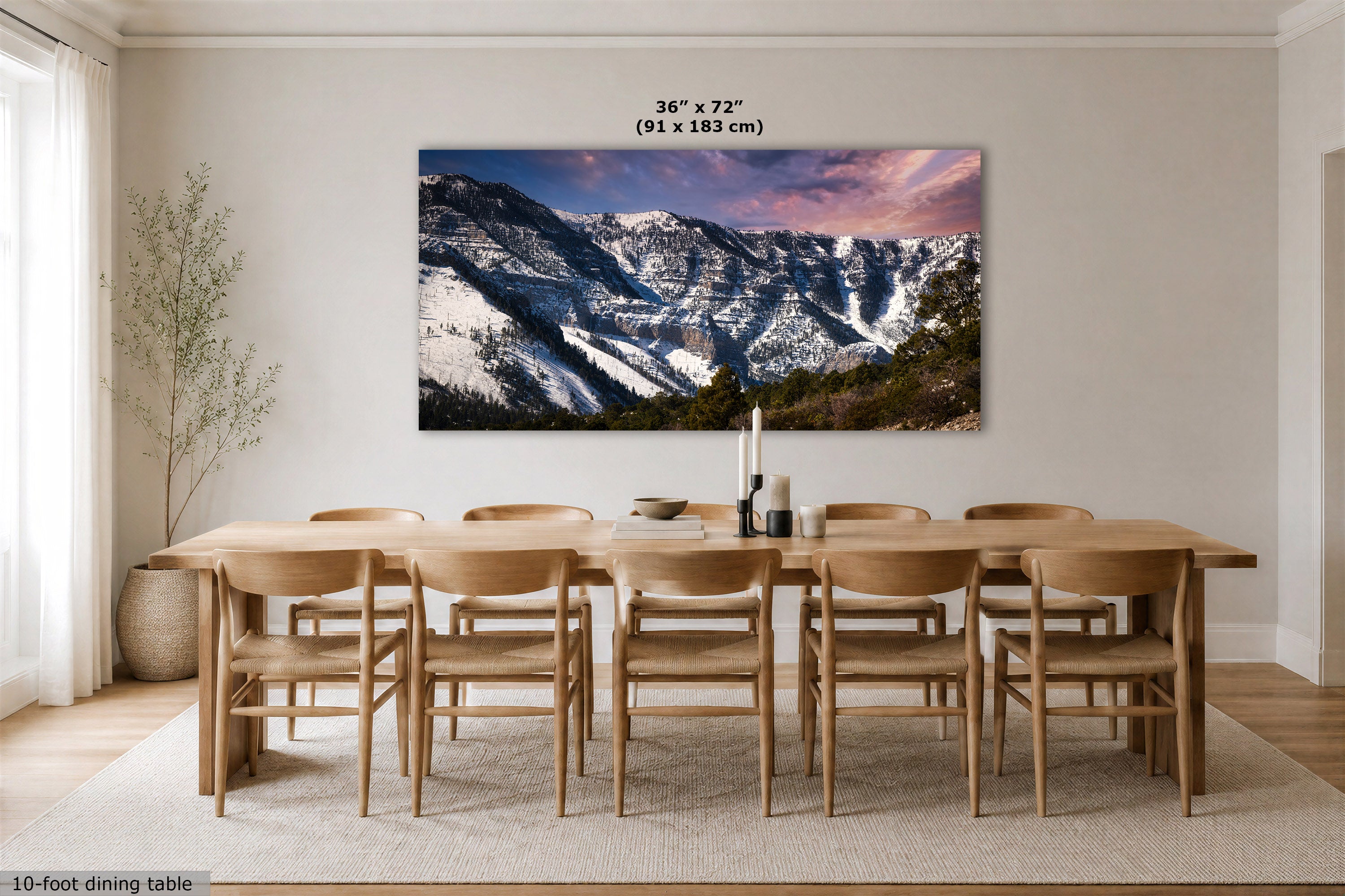A large rectangular wall art print showing snowy mountain peaks under a pink and purple sky, displayed above a wooden dining table and chairs in a bright dining room. Text above the print indicates dimensions 36 x 72 in (91 x 183 cm).