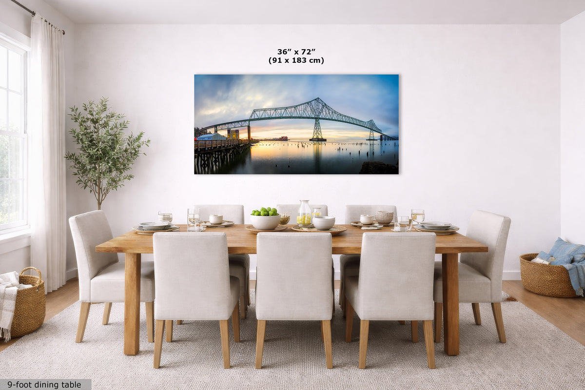 A framed wall art print of the Forth Bridge in Edinburgh, Scotland, displayed in a modern dining room setting above a wooden dining table and chairs. The print features a blue and white bridge over water at sunset, with dimensions '36" x 72" (91 x 183 cm)' printed above it.
