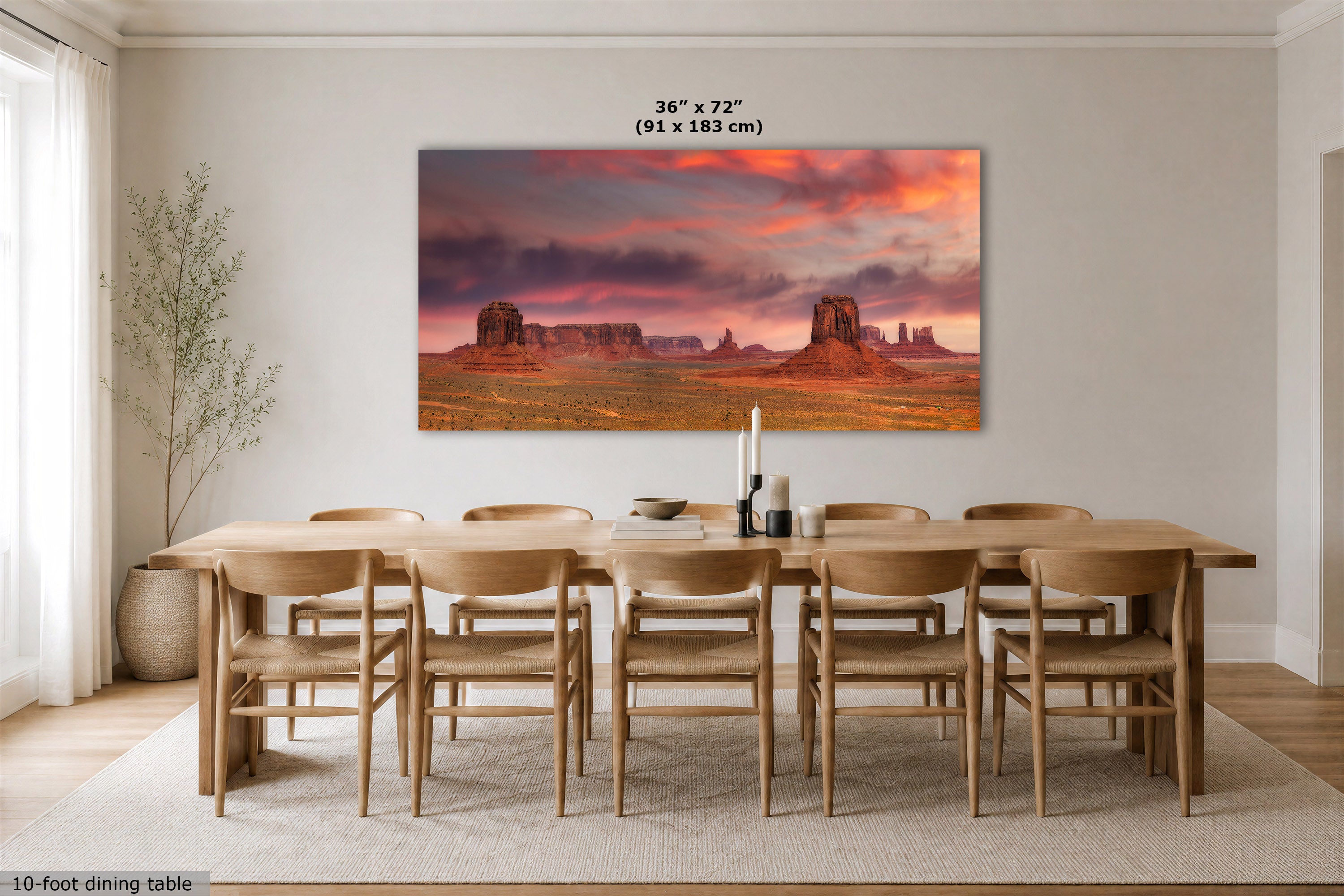 A large rectangular wall art print depicting a desert landscape at sunset hangs above a wooden dining table and chairs in a bright dining room. The print shows orange and pink clouds over red rock formations.
