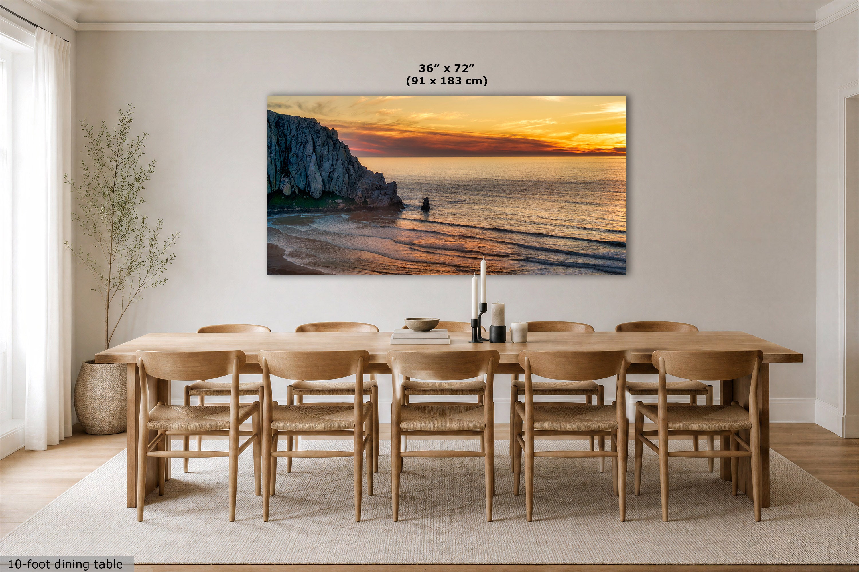 A large rectangular wall art print showing a coastal cliff and beach sunset hangs above a wooden dining table and chairs in a bright modern dining room. The print includes size text reading 36 x 72 (91 x 183 cm).